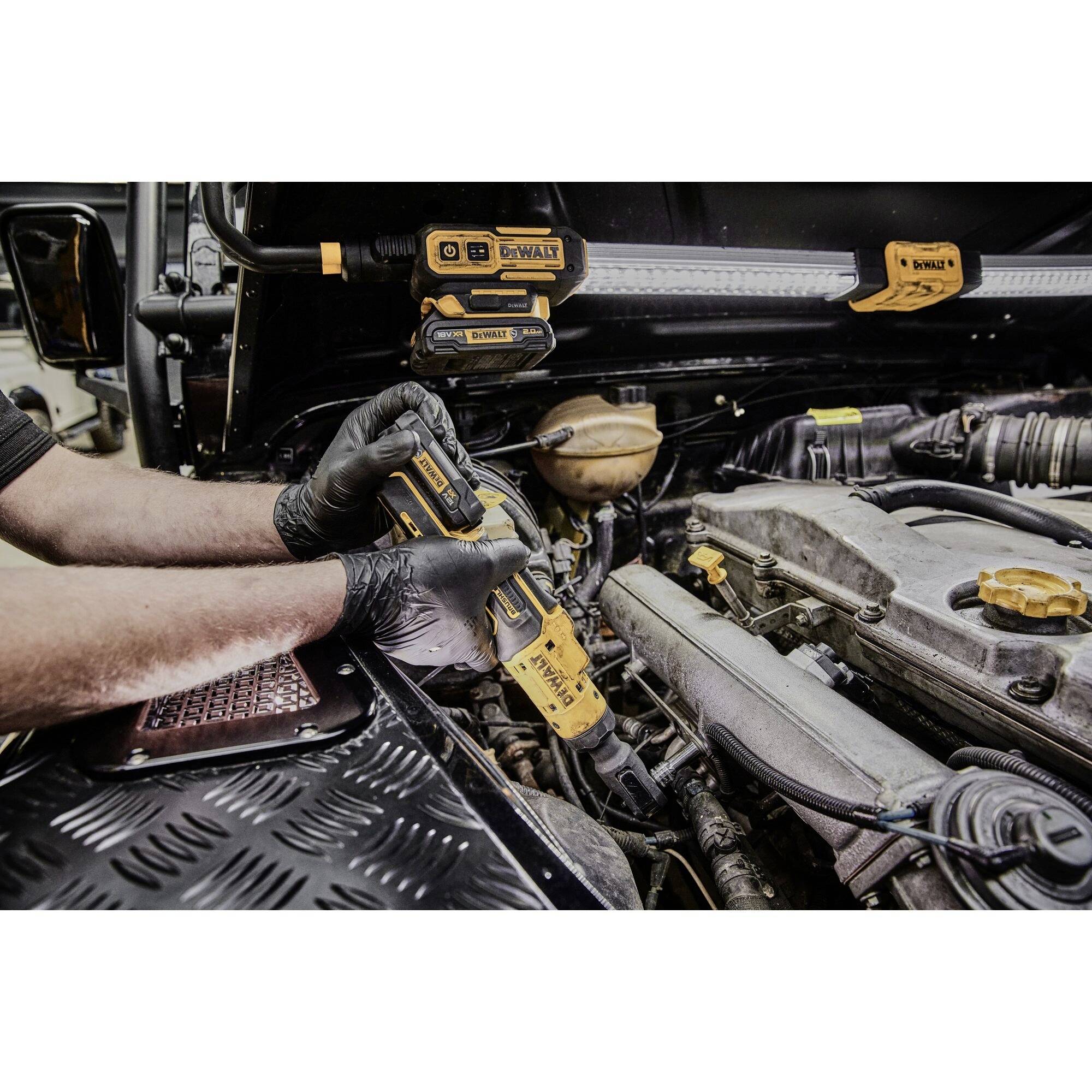 A person is working with a cordless drill in the engine compartment of a vehicle. Various tools and car parts are visible.