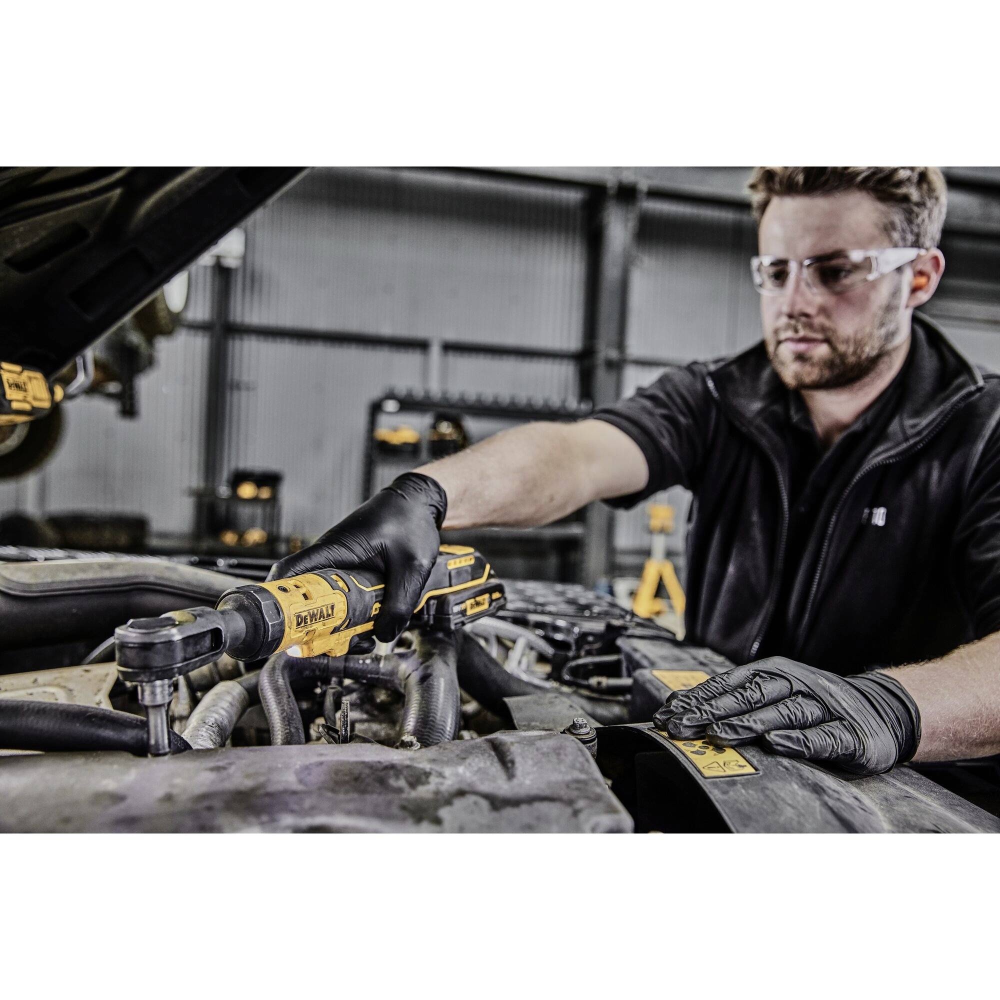 A man is working with a cordless drill on a car engine in a workshop. He is wearing safety glasses and gloves.