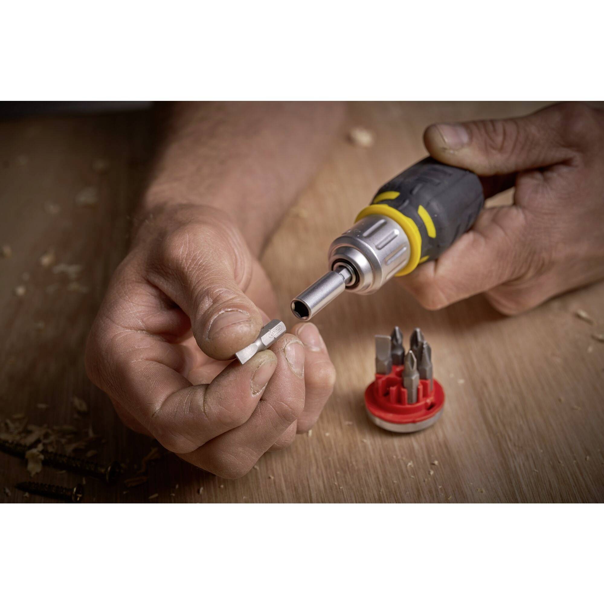 A hand is holding a screwdriver and a drill bit, with a set of various drill heads lying beside it on a wooden table.