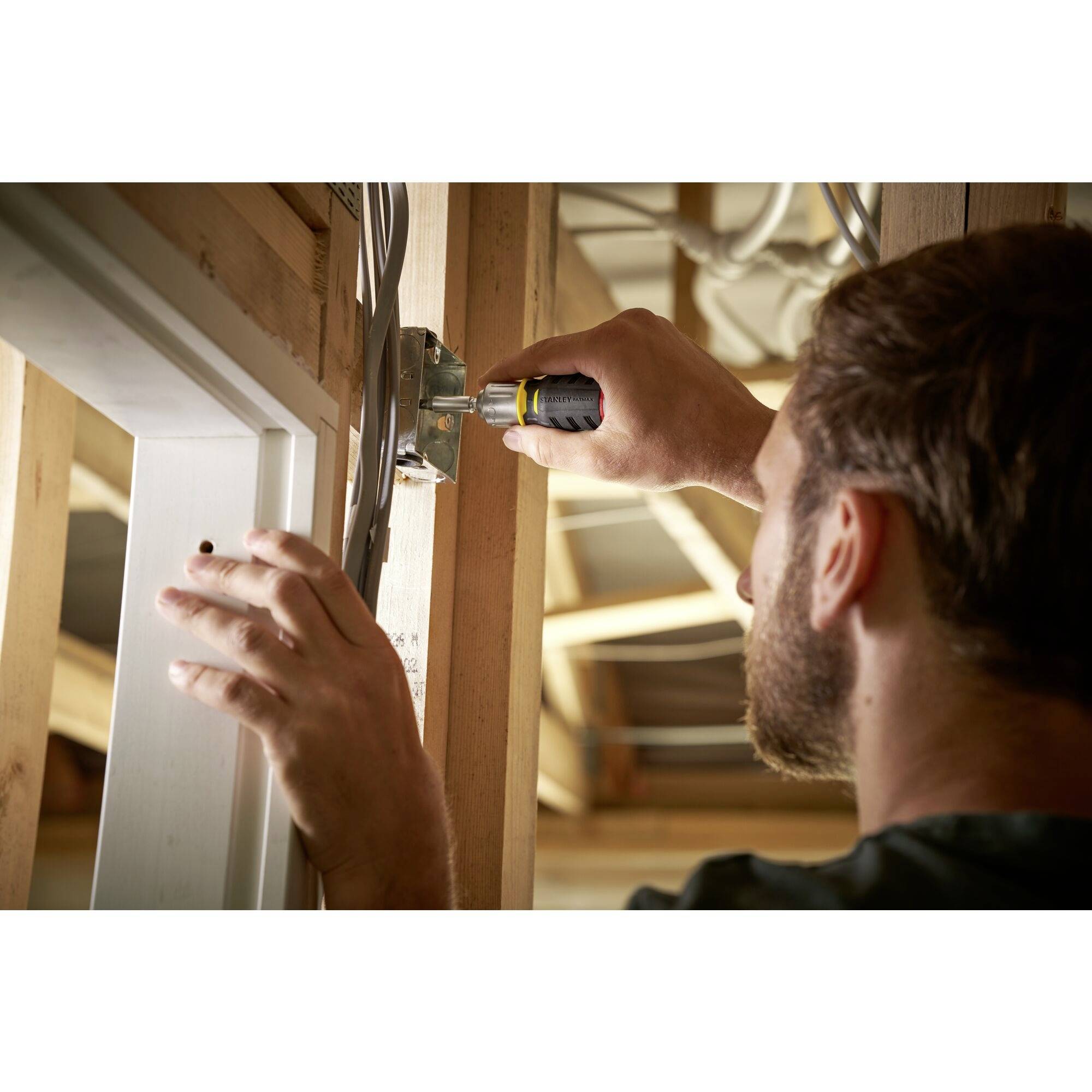 A person is securing an electrical plate to a wooden frame in an unfinished room using a screwdriver.