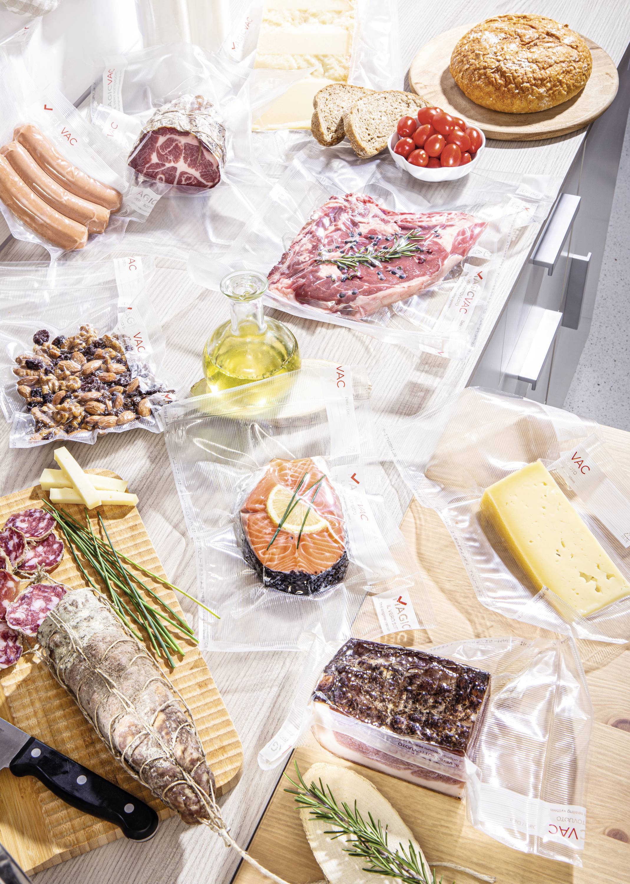 A kitchen worktop with vacuum-packed foods: sausage, meat, fish, cheese and bread. A loaf of bread, cherry tomatoes and oil are also visible.