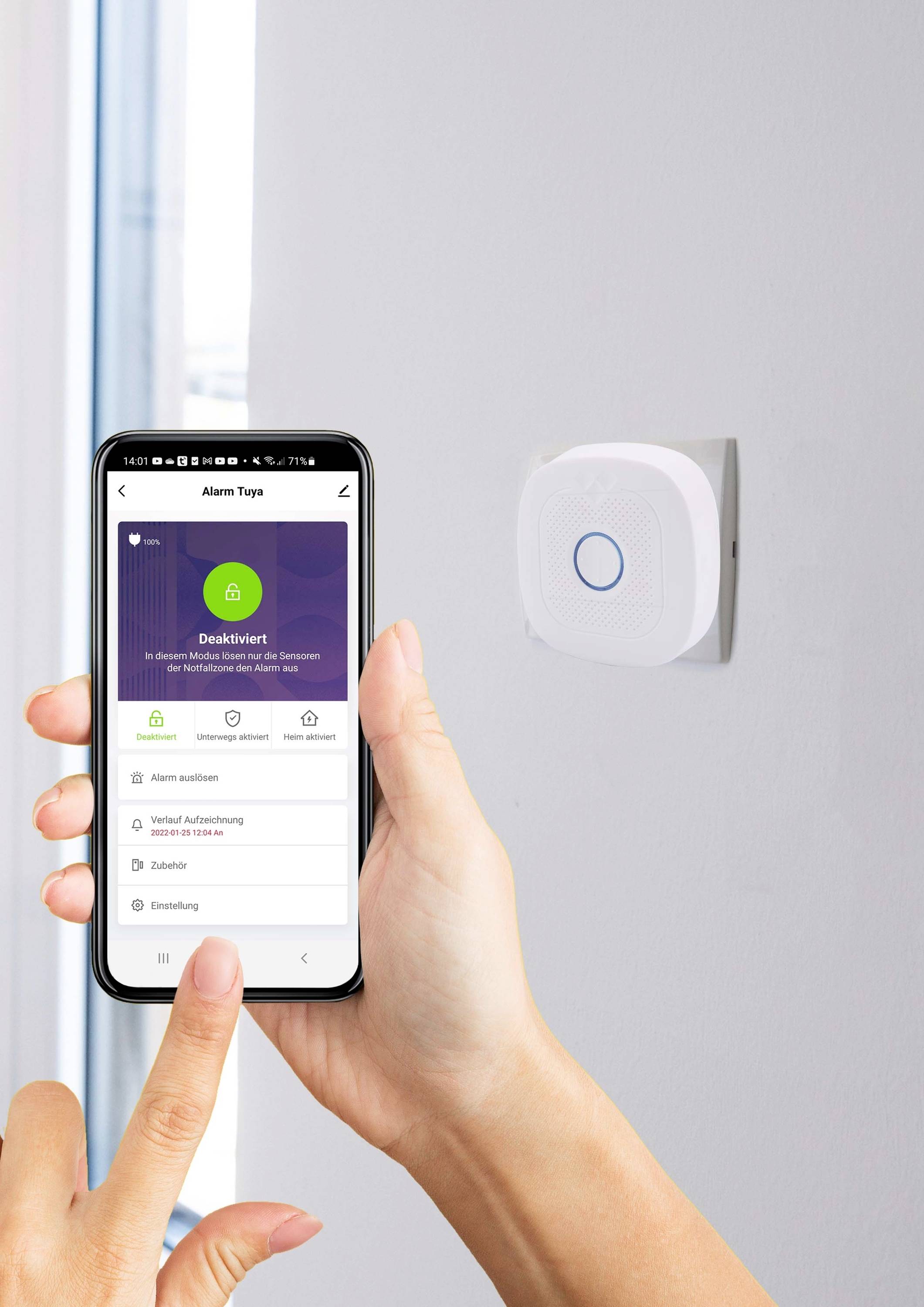 A person is holding a smartphone displaying an alarm app in front of a smoke detector mounted on the wall. A window is visible in the background.