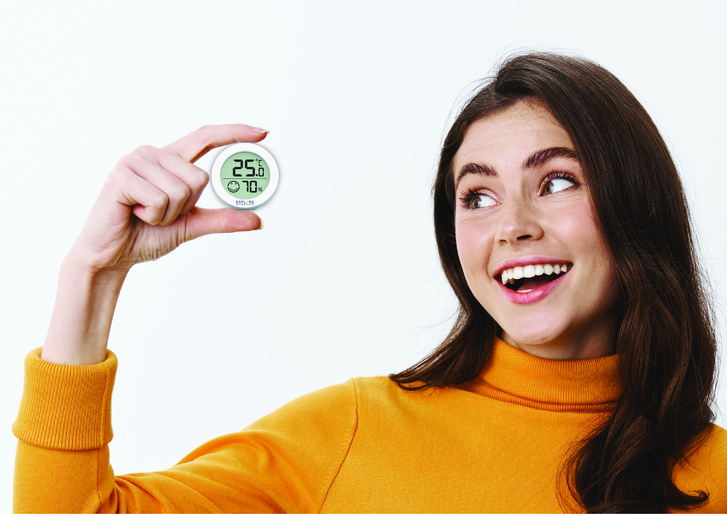 Woman in an orange jumper holding a round digital thermometer and hygrometer, showing 25.6°C and 70% humidity, smiling.