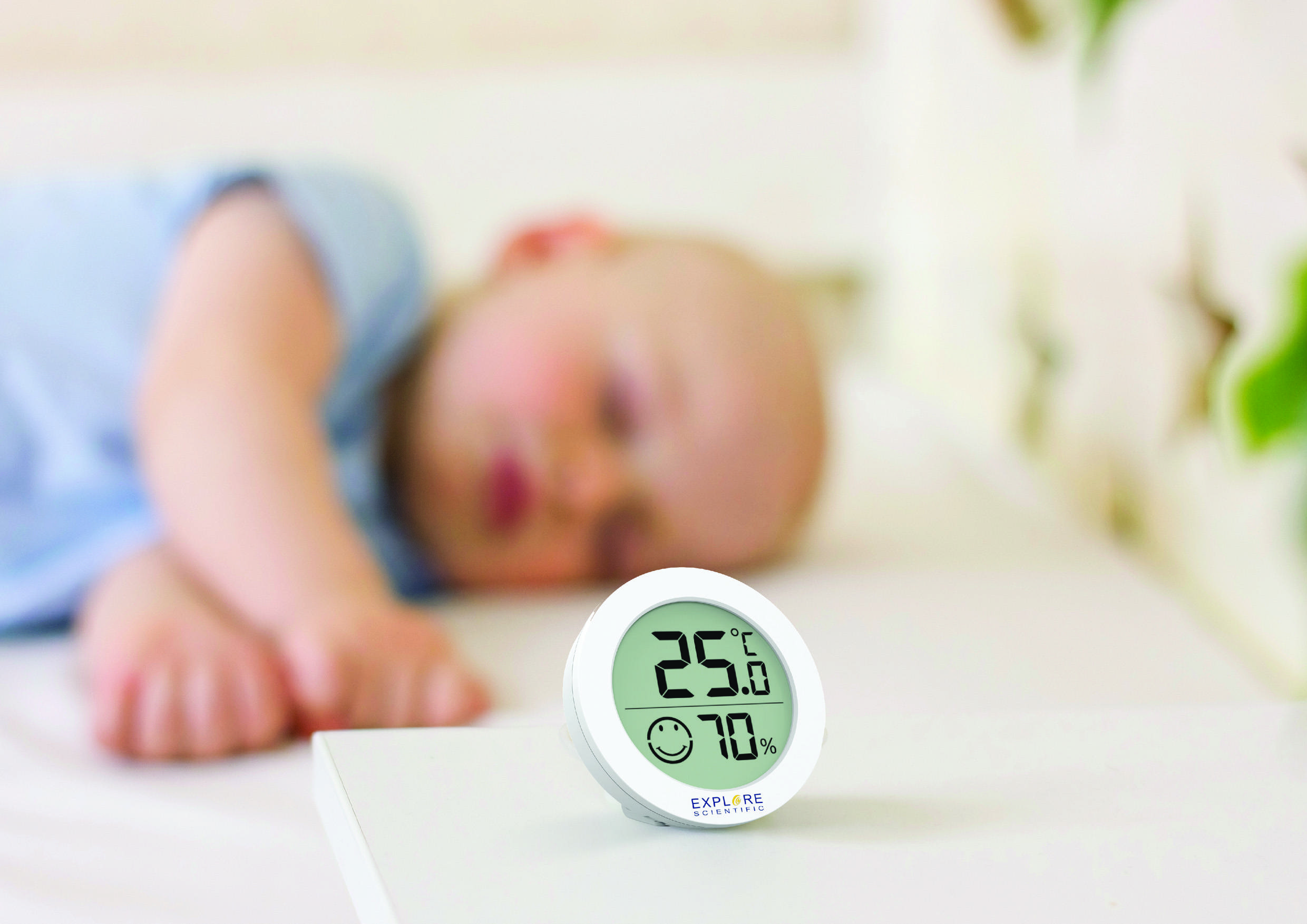 A sleeping baby in the background; in the foreground a digital thermo-hygrometer displaying 25°C and 70% relative humidity.