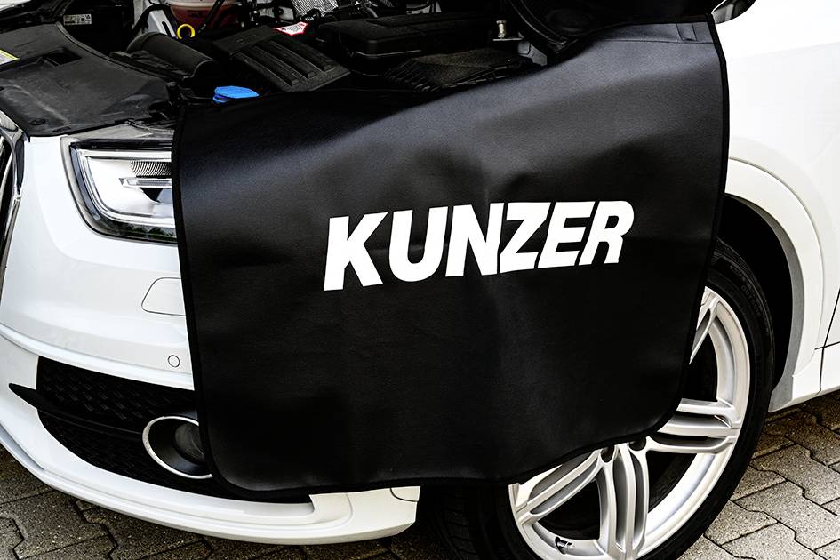 A white car with an open bonnet, covered with a black protective mat bearing the inscription 'KUNZER'.