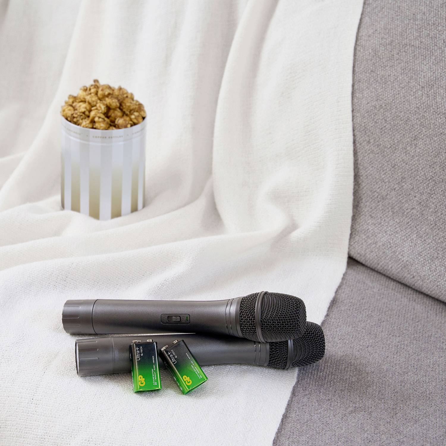 A black microphone and batteries are lying on a white blanket on a grey sofa. A tin of popcorn stands in the background.