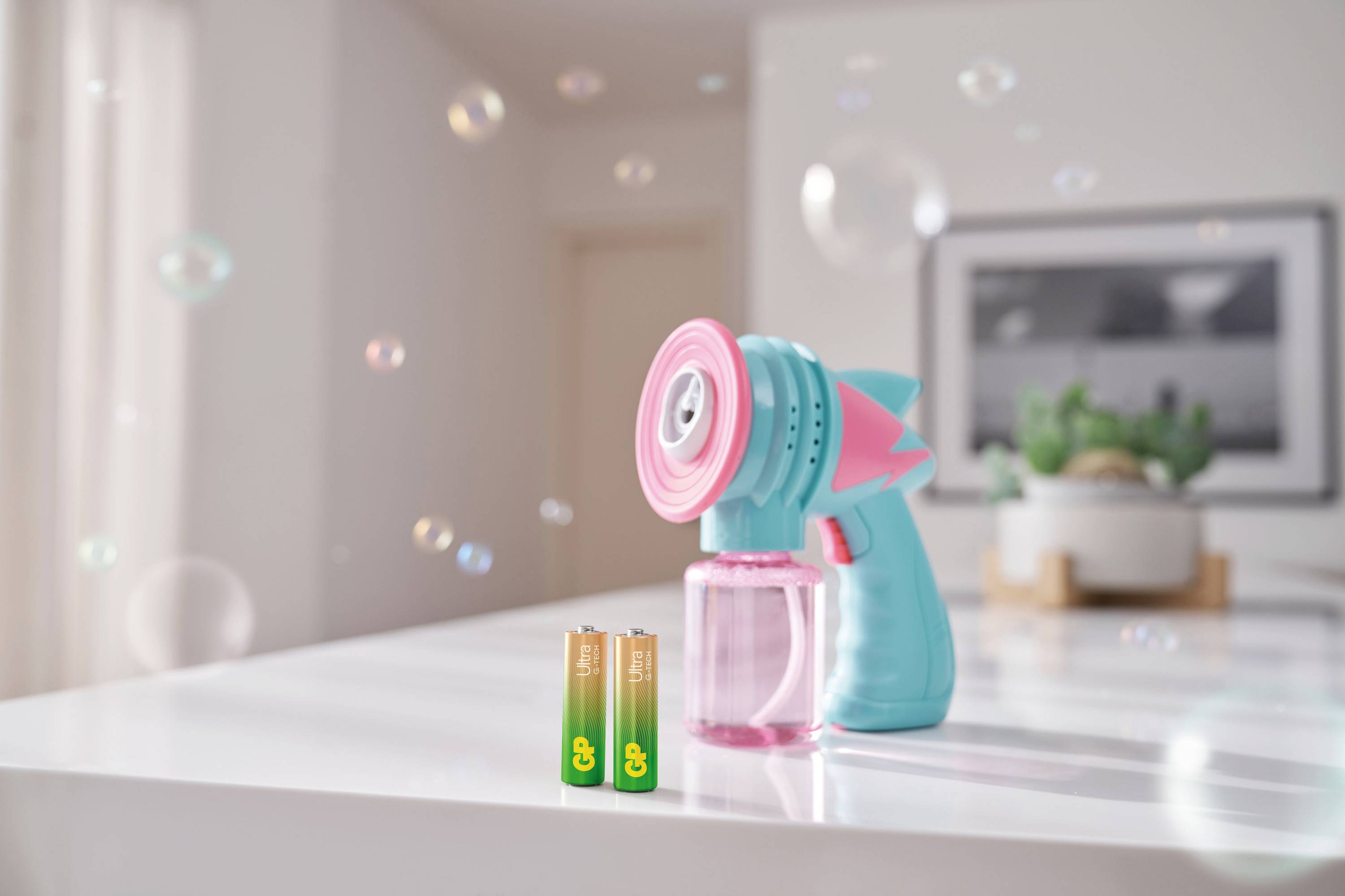 Soap bubble machine in the shape of a turquoise-pink gun blows bubbles; in front of it lie two AA batteries on a white table.