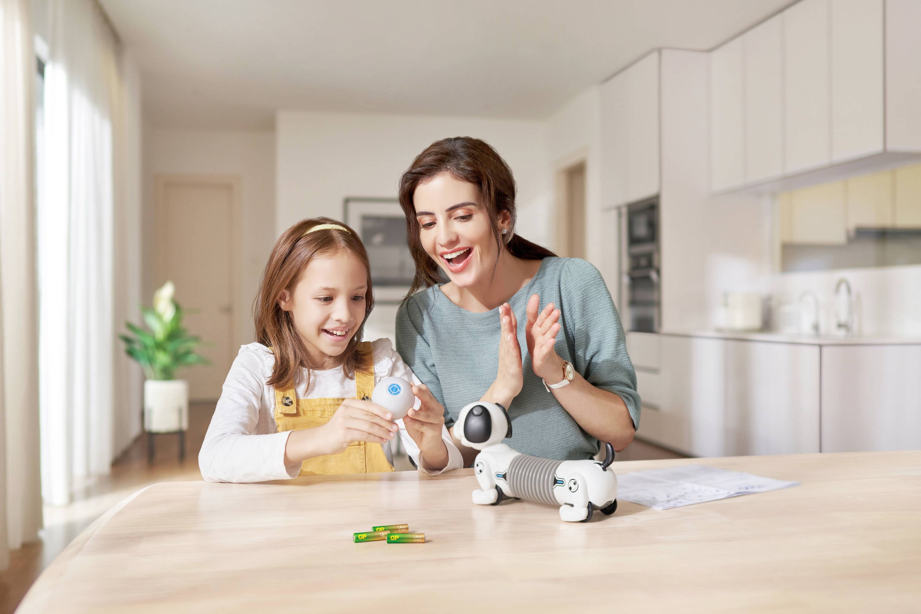 A woman and a girl jointly operate a small robot on the kitchen table in a bright flat.