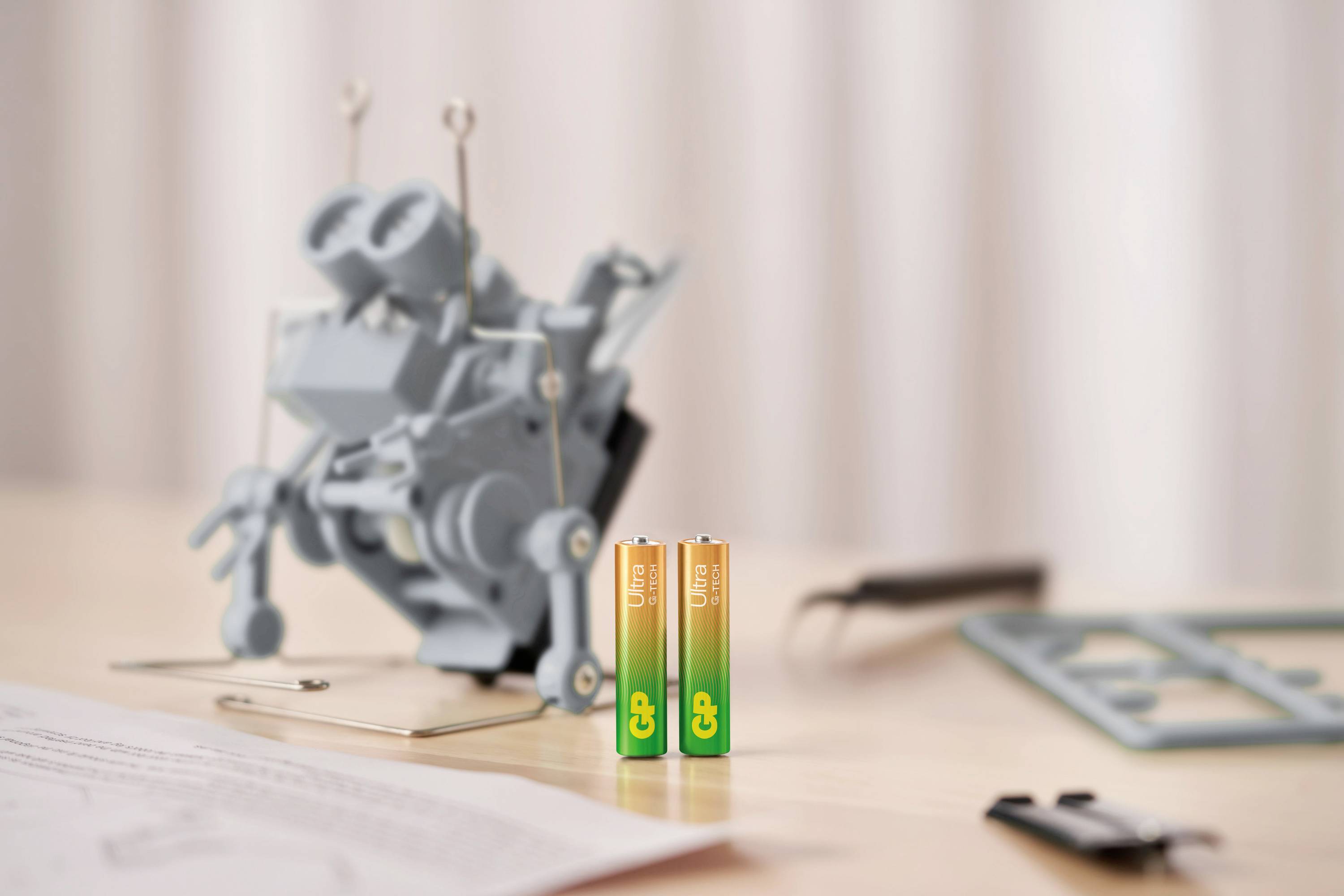 Two AA batteries in the foreground; behind them, slightly out of focus, a small robot on a table, with tools and parts lying to the right.