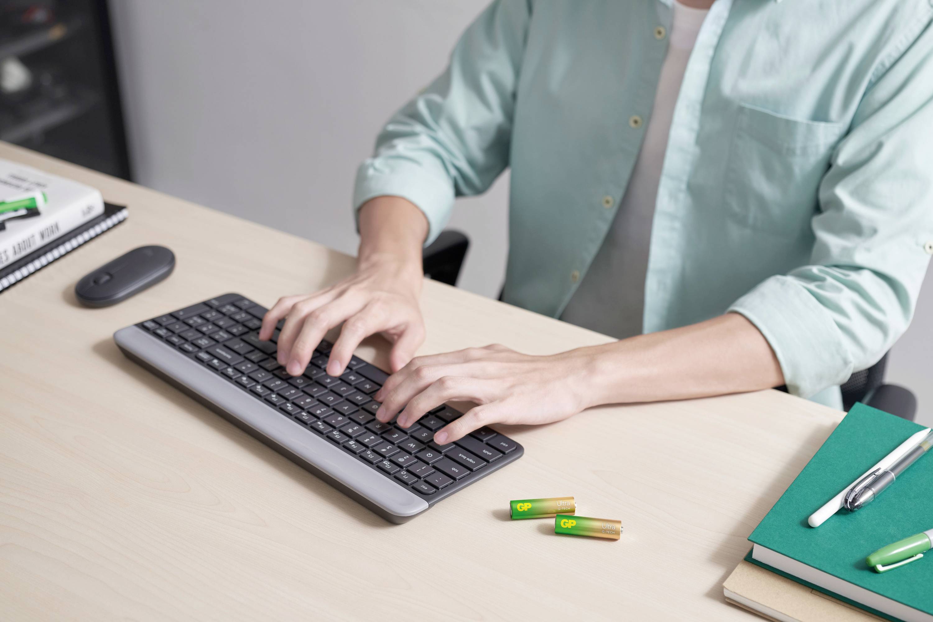 A person is typing with both hands on a keyboard at a desk; next to it are batteries, a mouse, a pen and notebooks.
