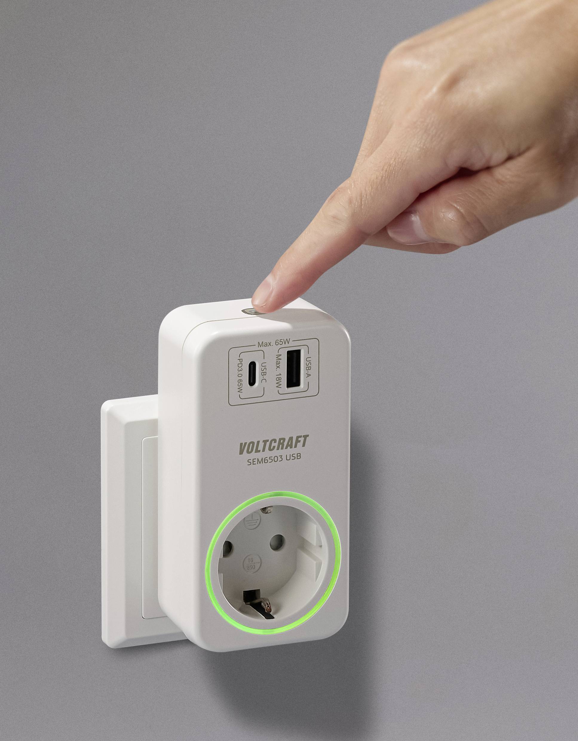 A hand is pointing at a white plug adapter with USB ports and a green illuminated ring indicator, mounted on a wall.