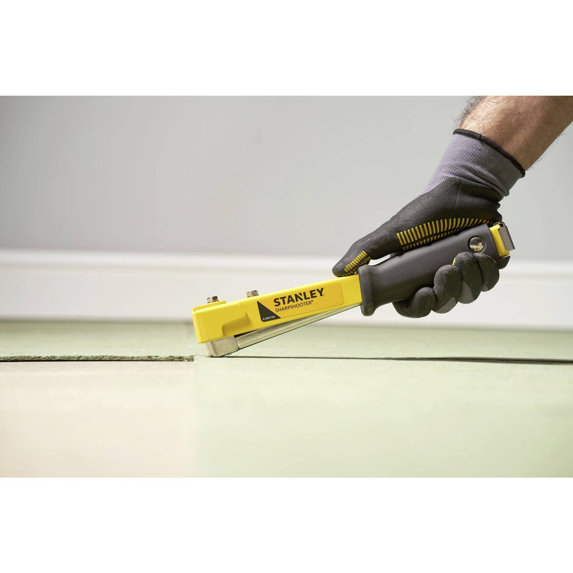 A gloved hand holds a yellow Stanley hand stapler that is being secured to the floor. The room has a light grey wall.