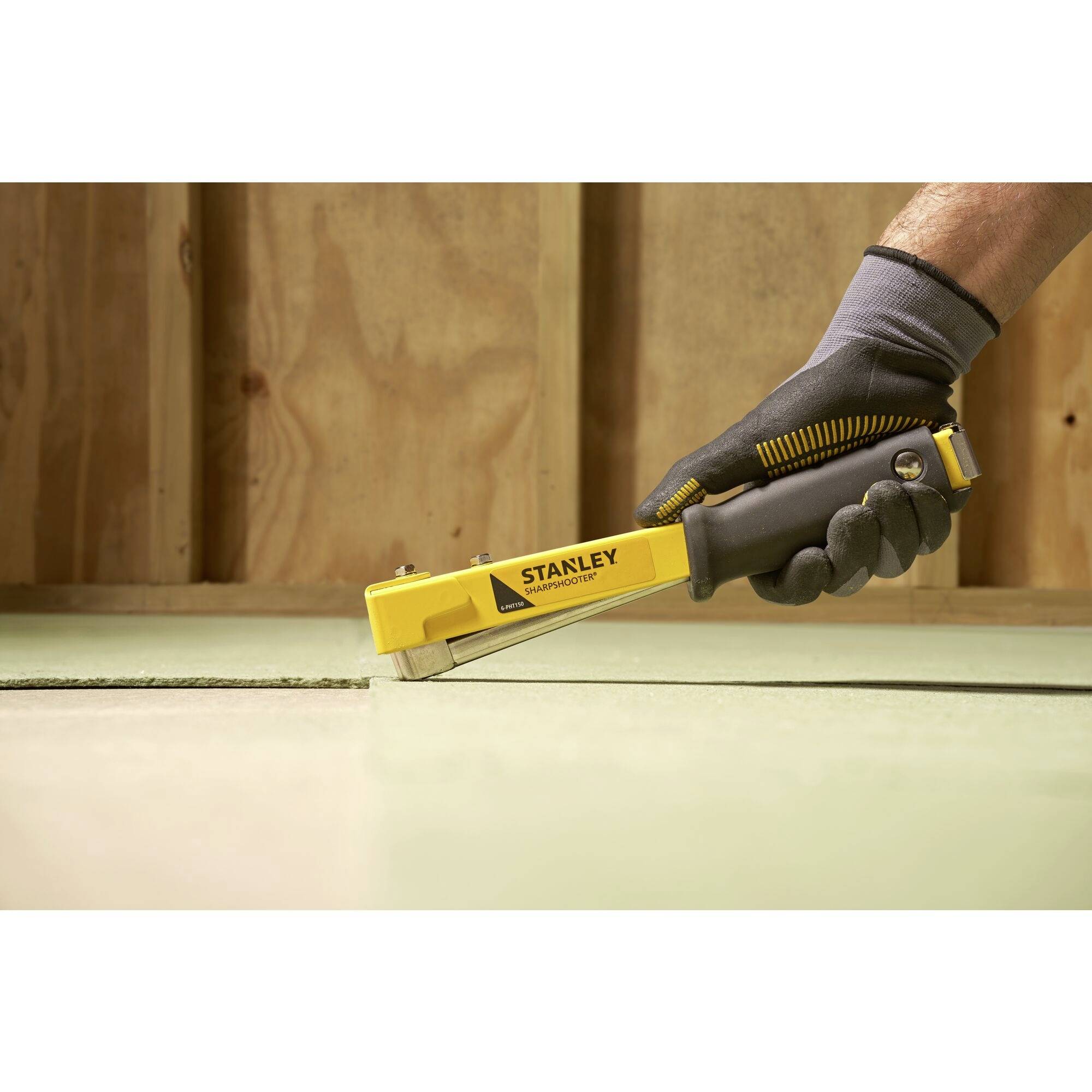 Close-up of a gloved hand operating a yellow Stanley staple gun. The staple gun is being used to secure material to wood.