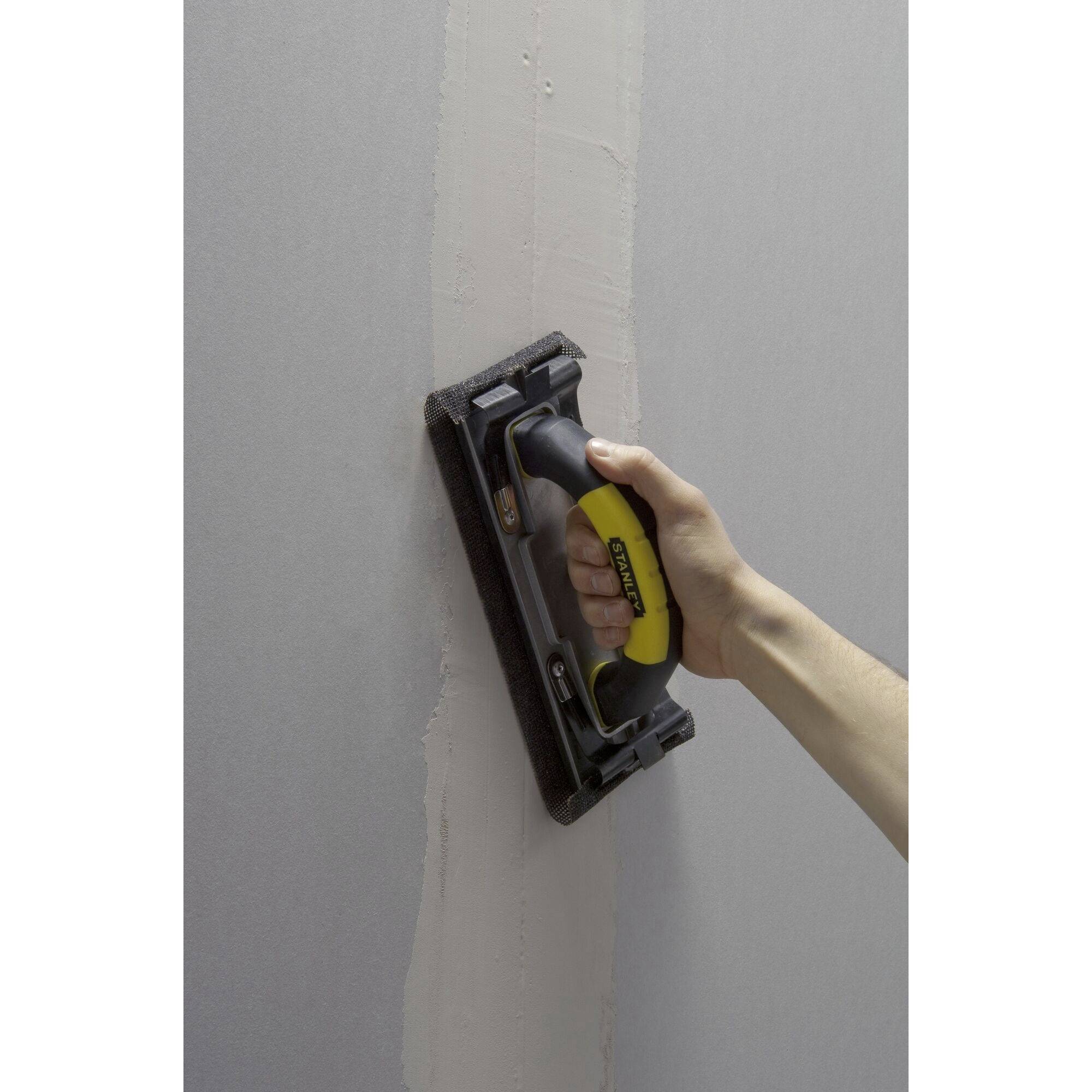 Hand sands plasterboard wall with sanding block and yellow handle.