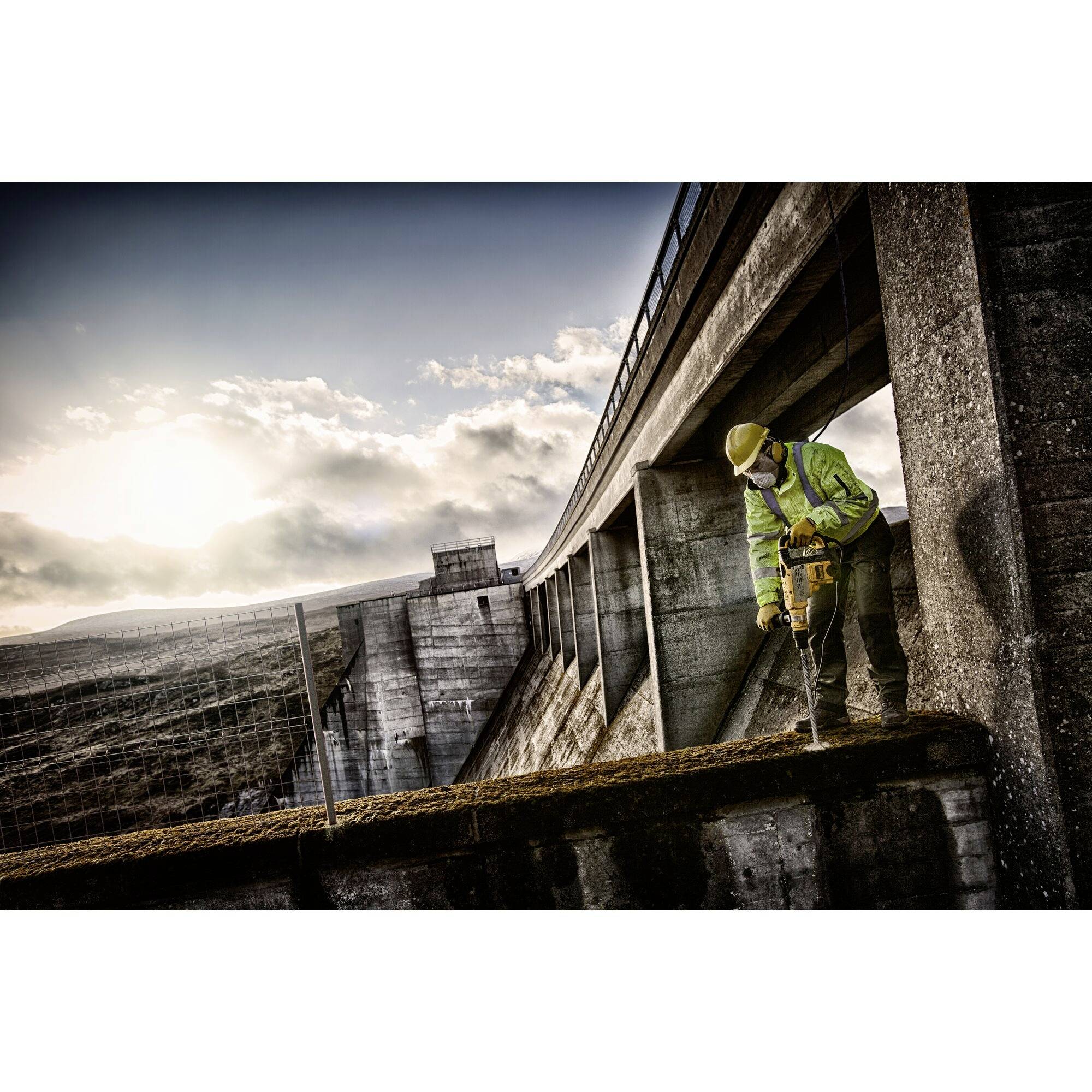 A construction worker in safety clothing works with a drill on a concrete structure outdoors, with the sun standing in the background.