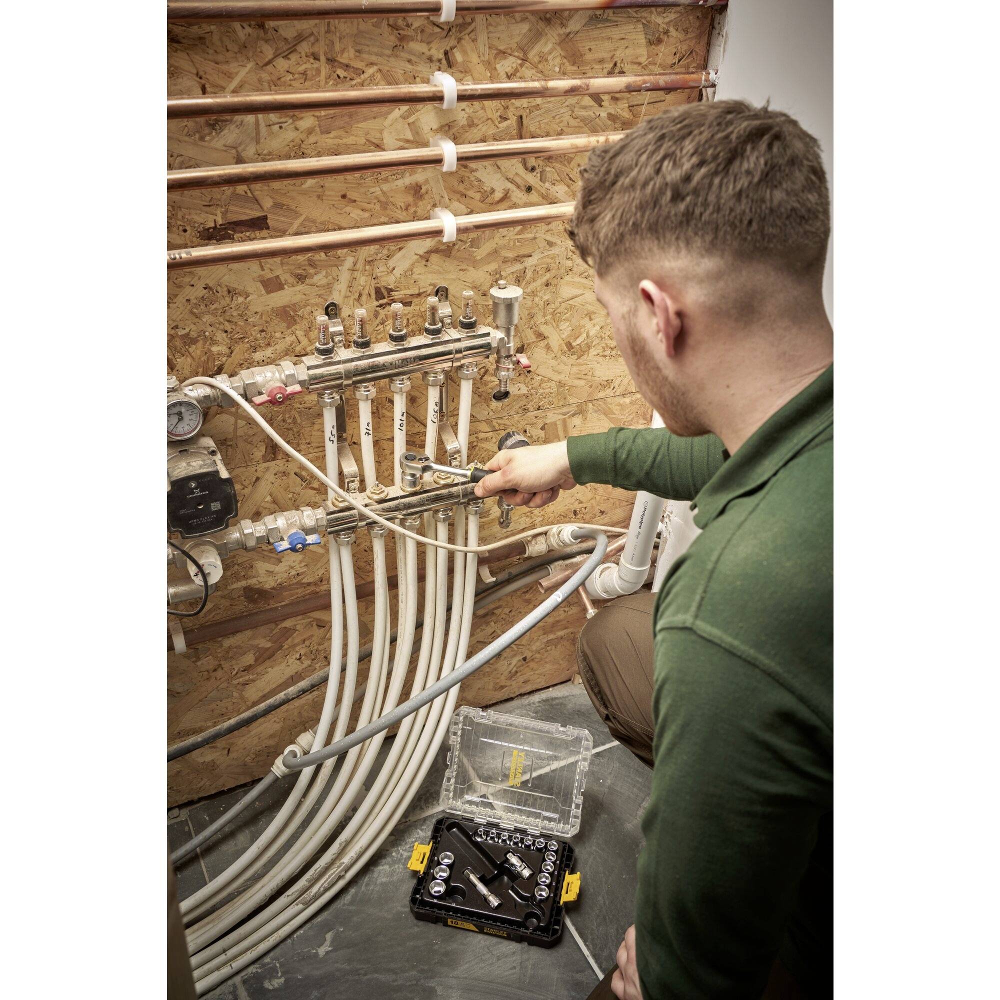 A man is working on a heating manifold with multiple pipes in a plant room. He is using a tool for adjustments.