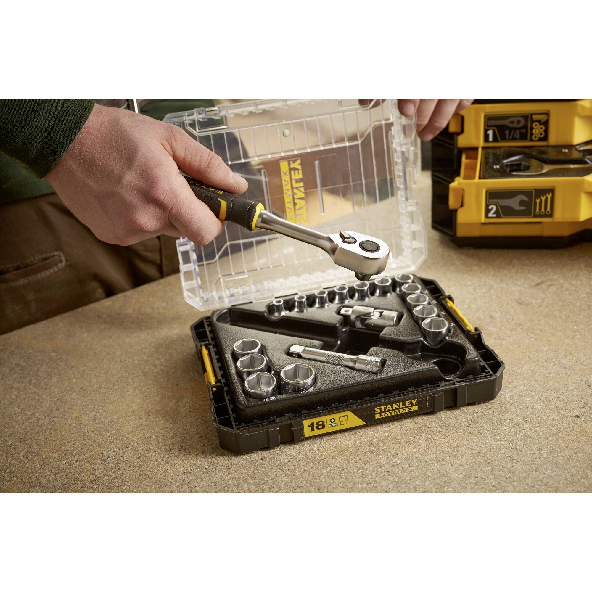 A person is holding a ratchet over an open socket set lying on a table. The set contains various sockets.