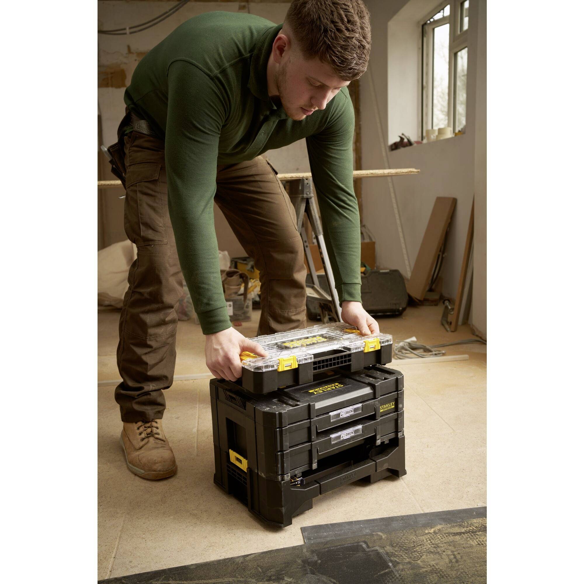 A man in workwear is stacking tool cases in a workshop. He is placing a transparent case on top of a larger black case.