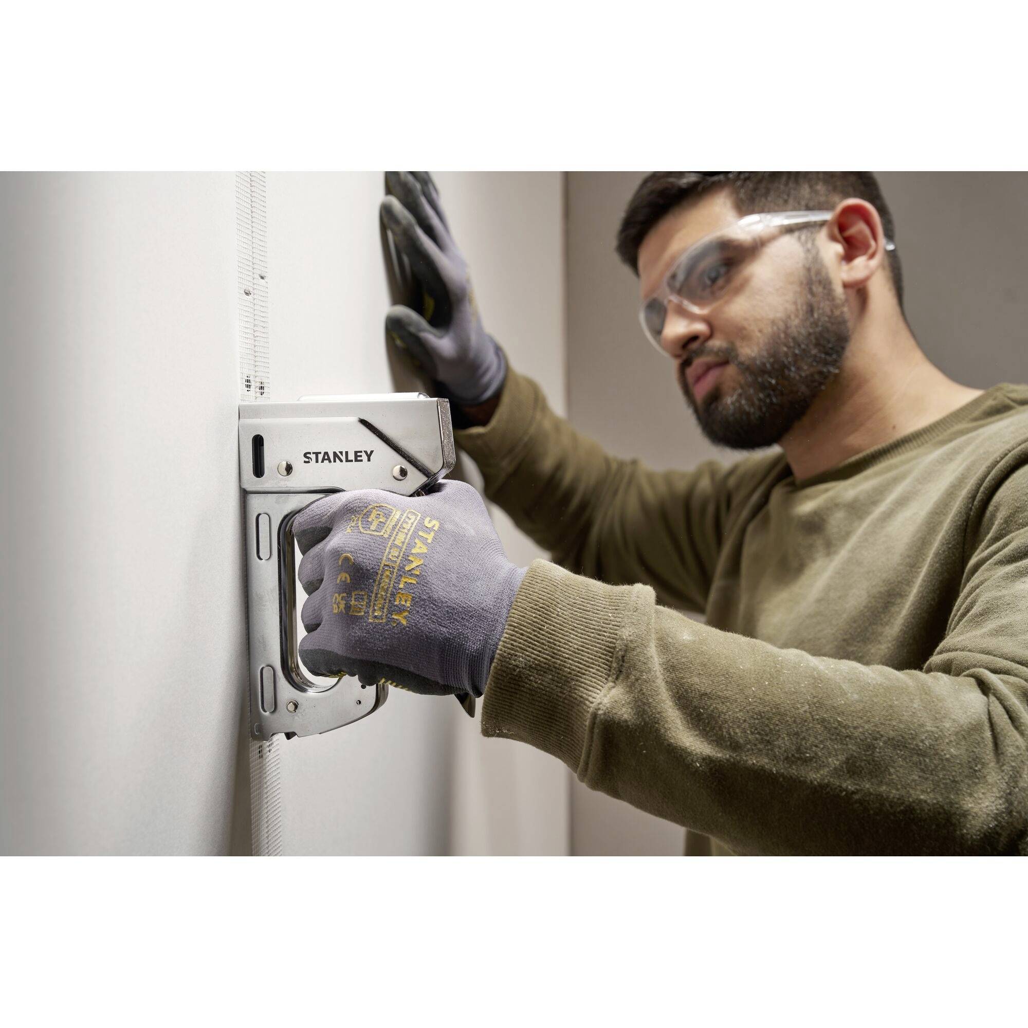 A person wearing safety goggles and gloves is securing a plasterboard sheet to the wall with a staple gun.