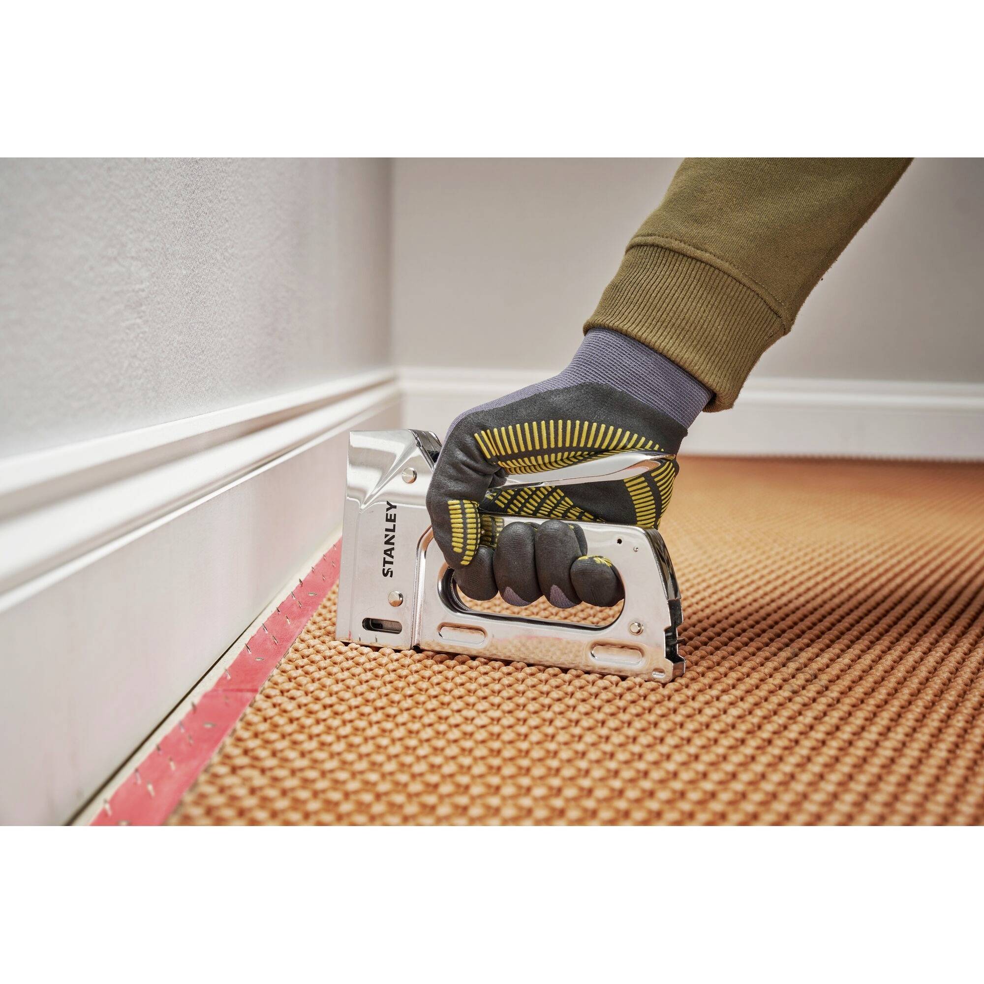 A gloved hand uses a staple gun to secure an orange carpet along a white skirting board.