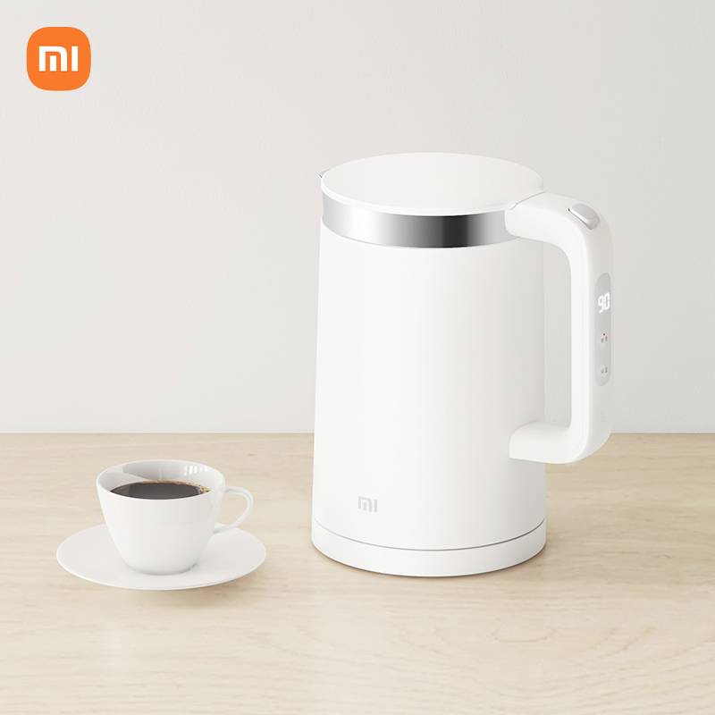 A white electric kettle is sitting on a table next to a cup of coffee. The Xiaomi logo is in the top left corner.