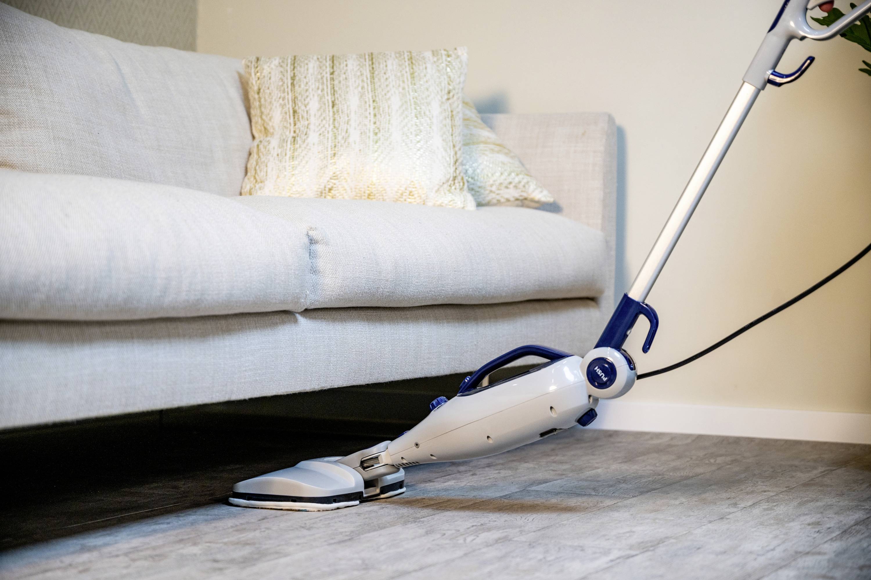 A steam cleaner is cleaning the floor underneath a white sofa on a wooden floor in a living room.