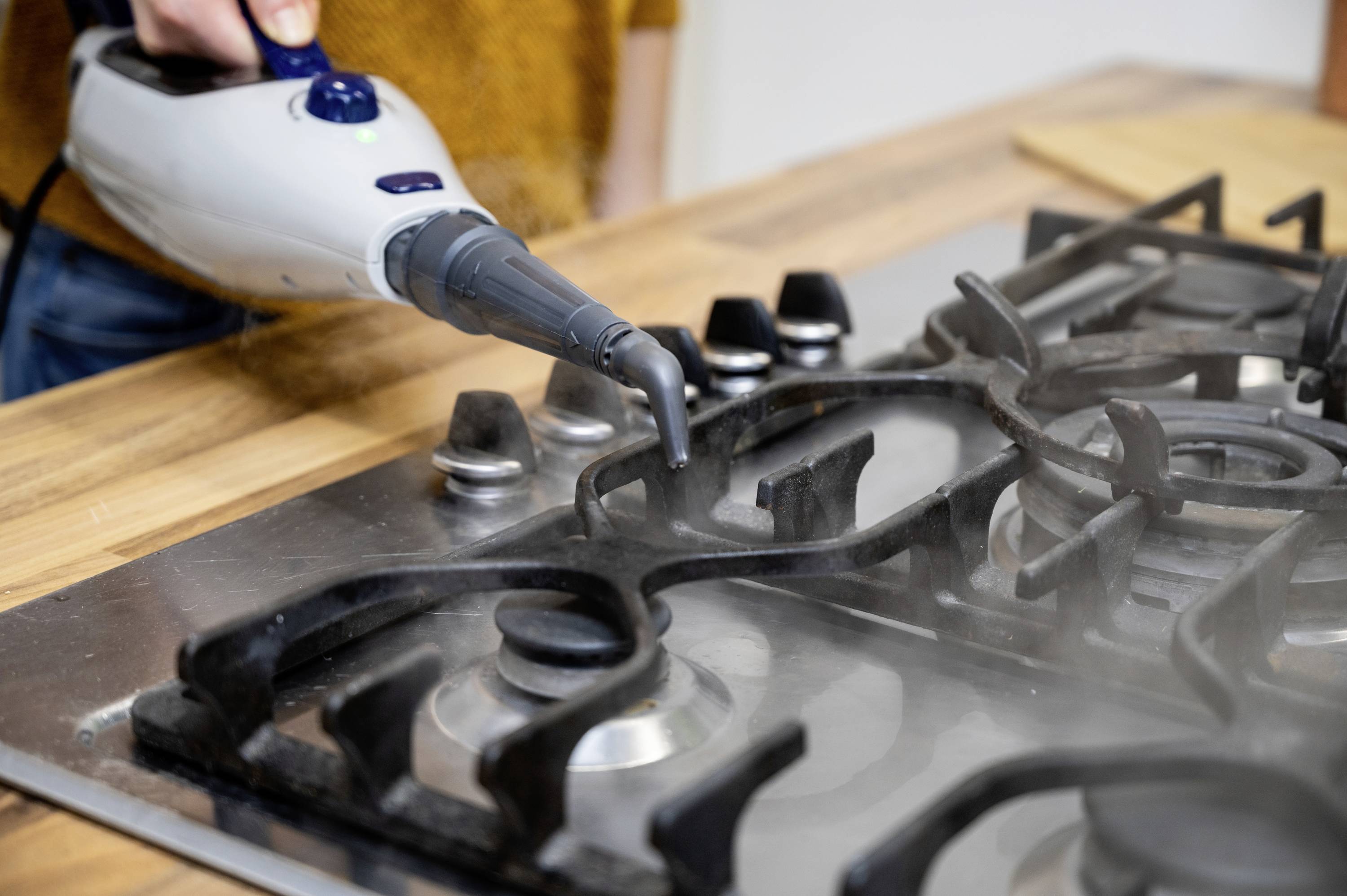 A person is cleaning a gas cooker with a steam cleaner. Steam rises as dirt is removed.