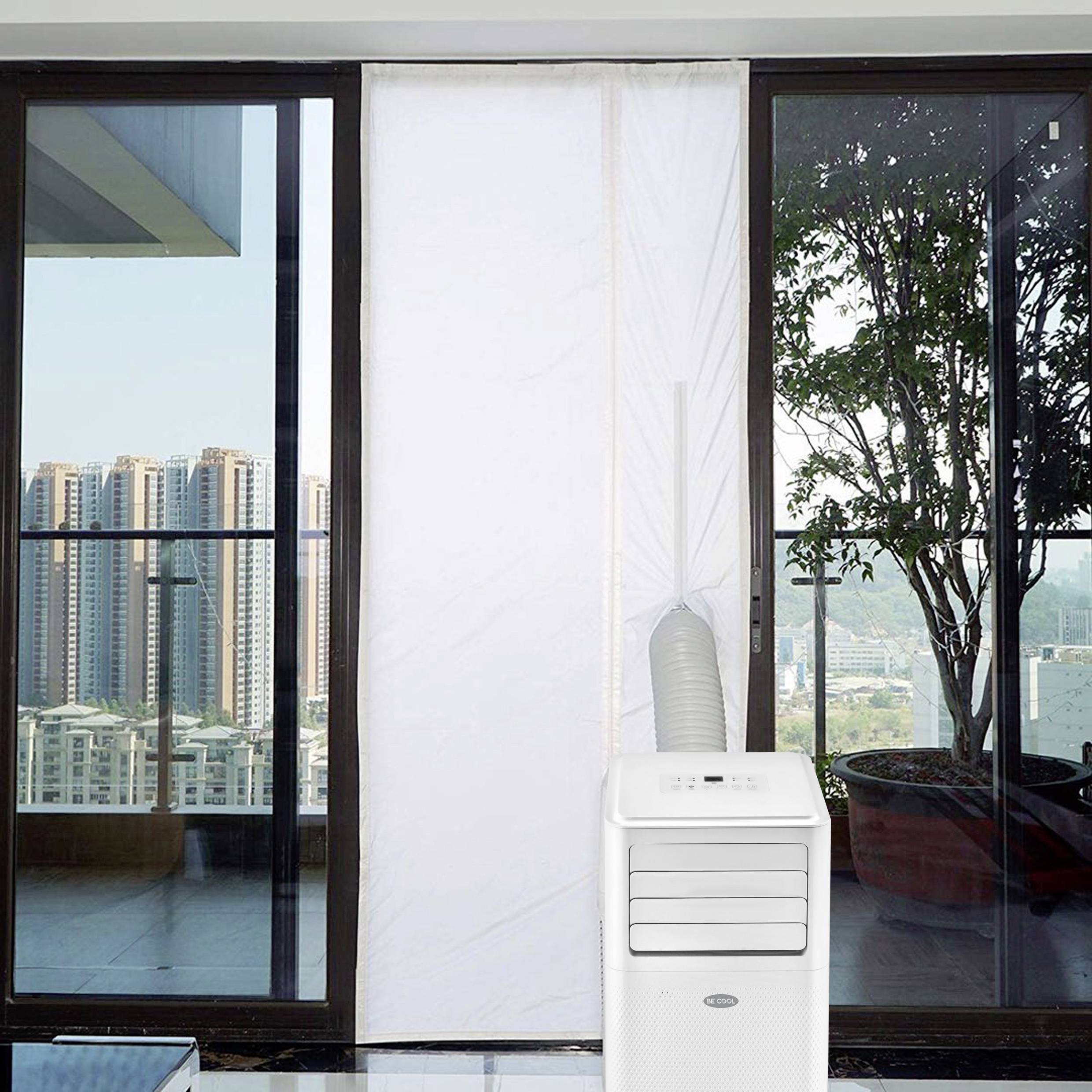 A mobile air cooler stands in front of a large window frontage. Outside, high-rise buildings and a sunny sky can be seen.