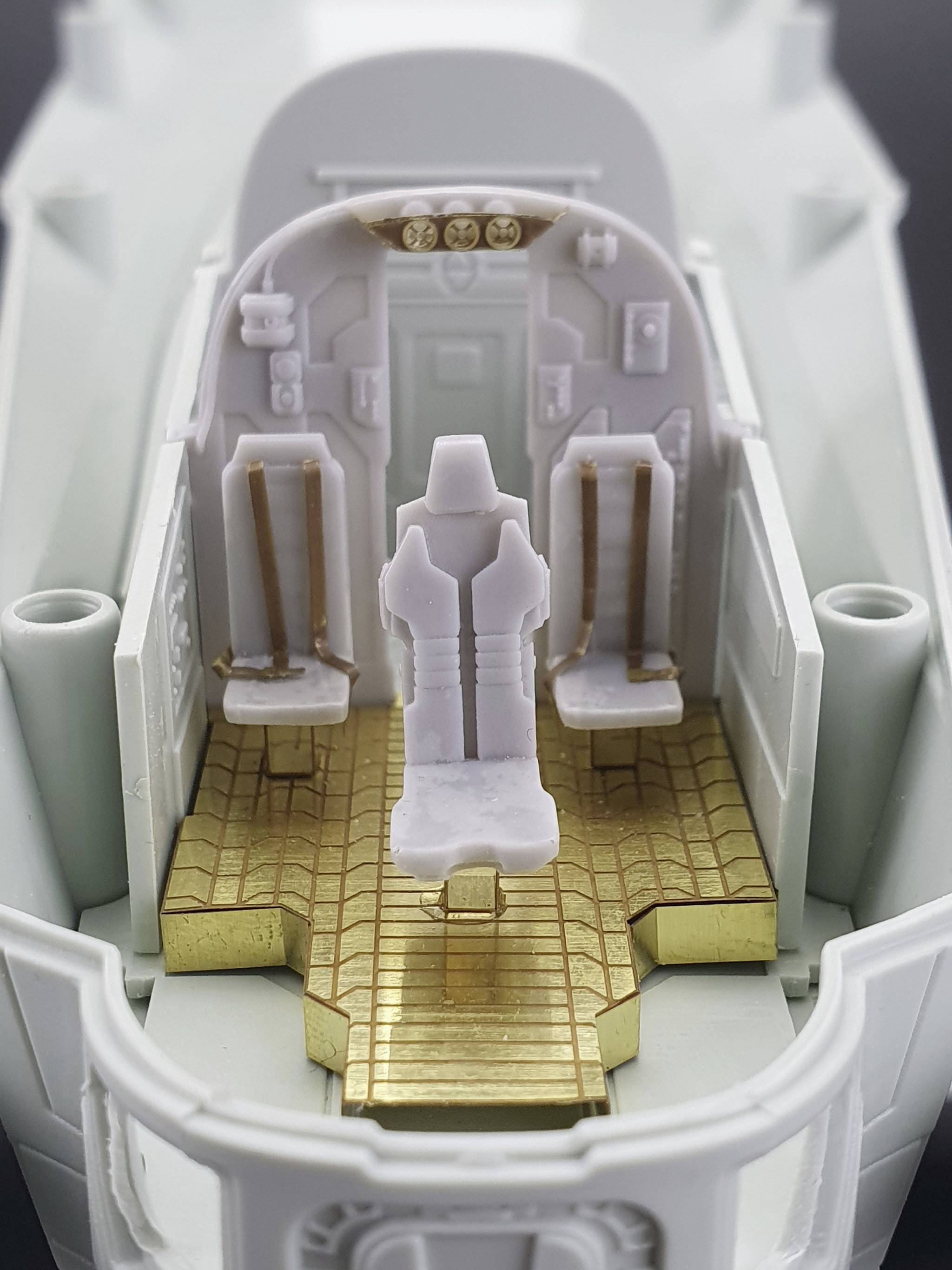 Model of a cockpit with three white seats and a gold-coloured floor. It shows detailed safety harnesses and instrument panel.