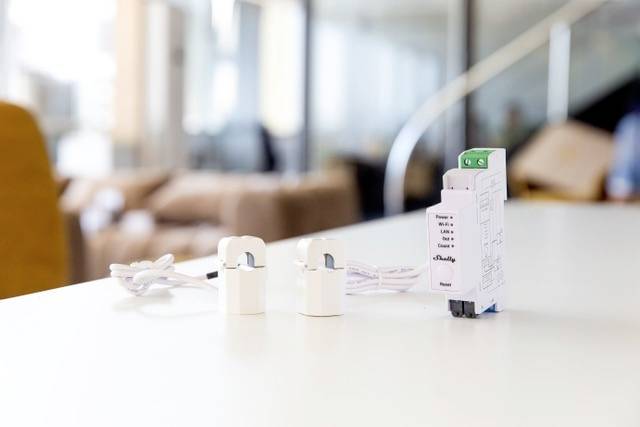 An energy measurement device with cables lies on a table in a modern office. A sofa is blurrily visible in the background.