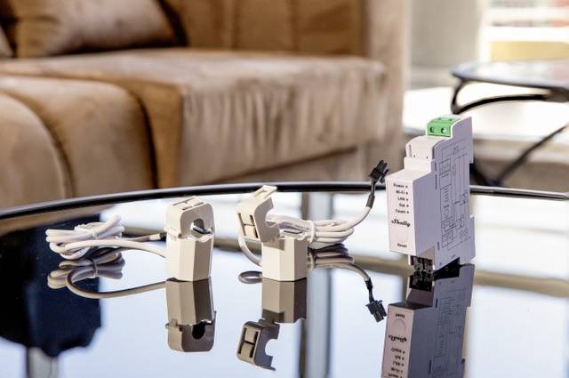 On a glass table, two white electronic devices with cables are lying, reflected on the table surface. A sofa is visible in the background.