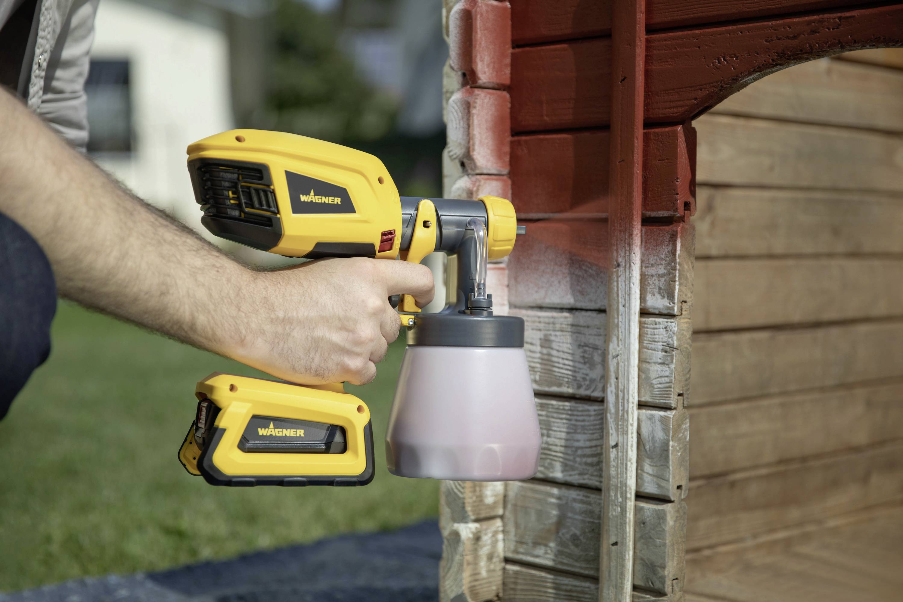 A person is applying red lacquer to a wooden surface using a yellow spray can. The setting is a garden.