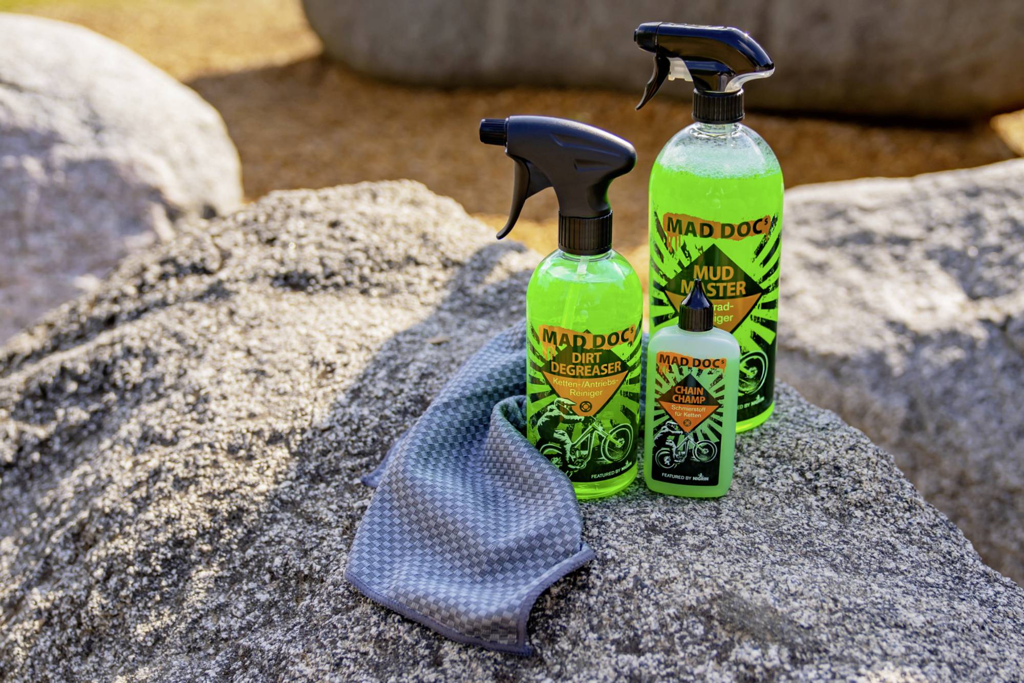 Three bottles of MAD DOG cleaning products and a cleaning cloth on a rock. Included are Dirt Degreaser and Mud Buster.