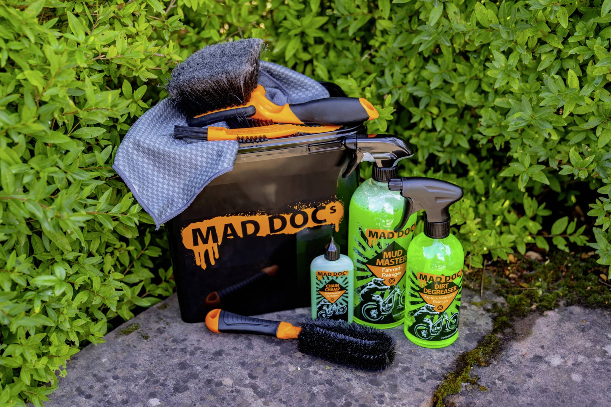 'Mad Doc's' cleaning set consisting of a black bucket, various brushes, and green bottles, placed on a stone in front of green bushes.