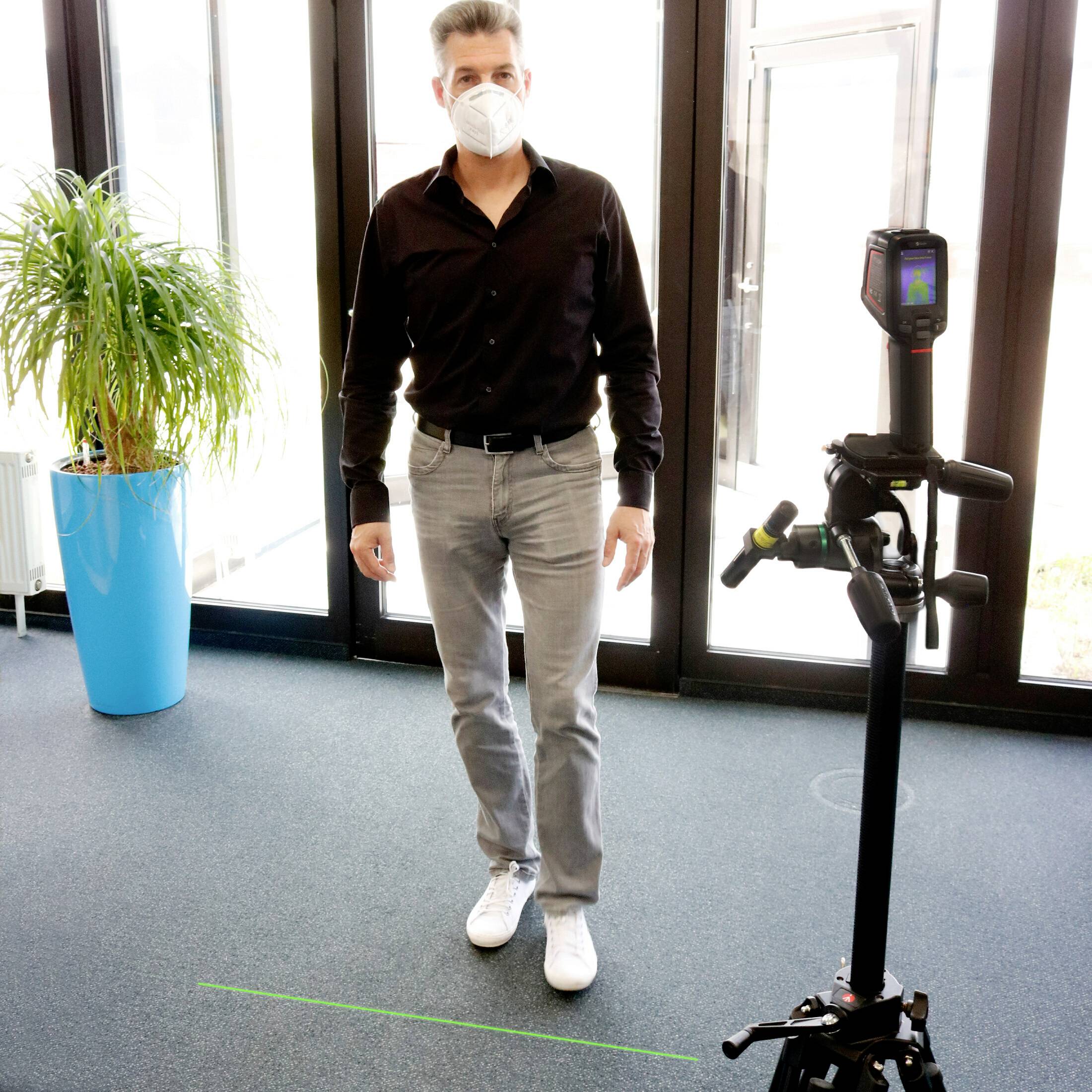 A person wearing a face mask is standing next to a potted plant and behind a measuring device in a bright room with glass doors.