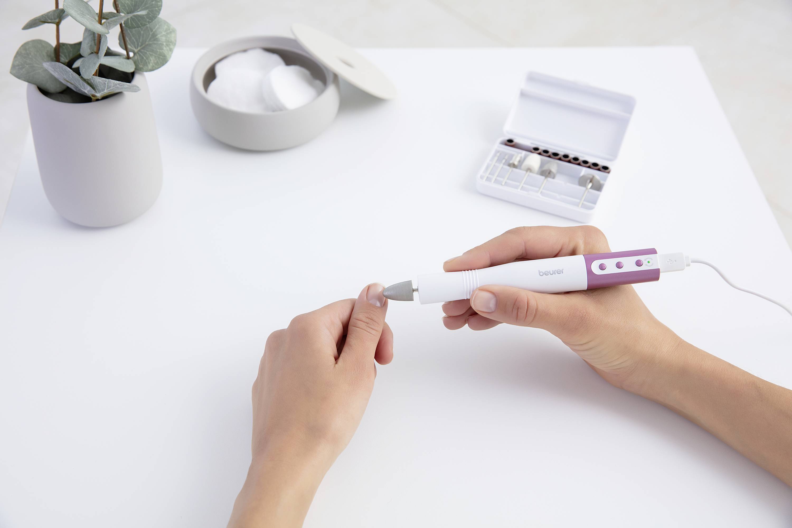 A person is manicuring their nails with an electric nail care device. Next to them, an open set with attachments sits on a table.