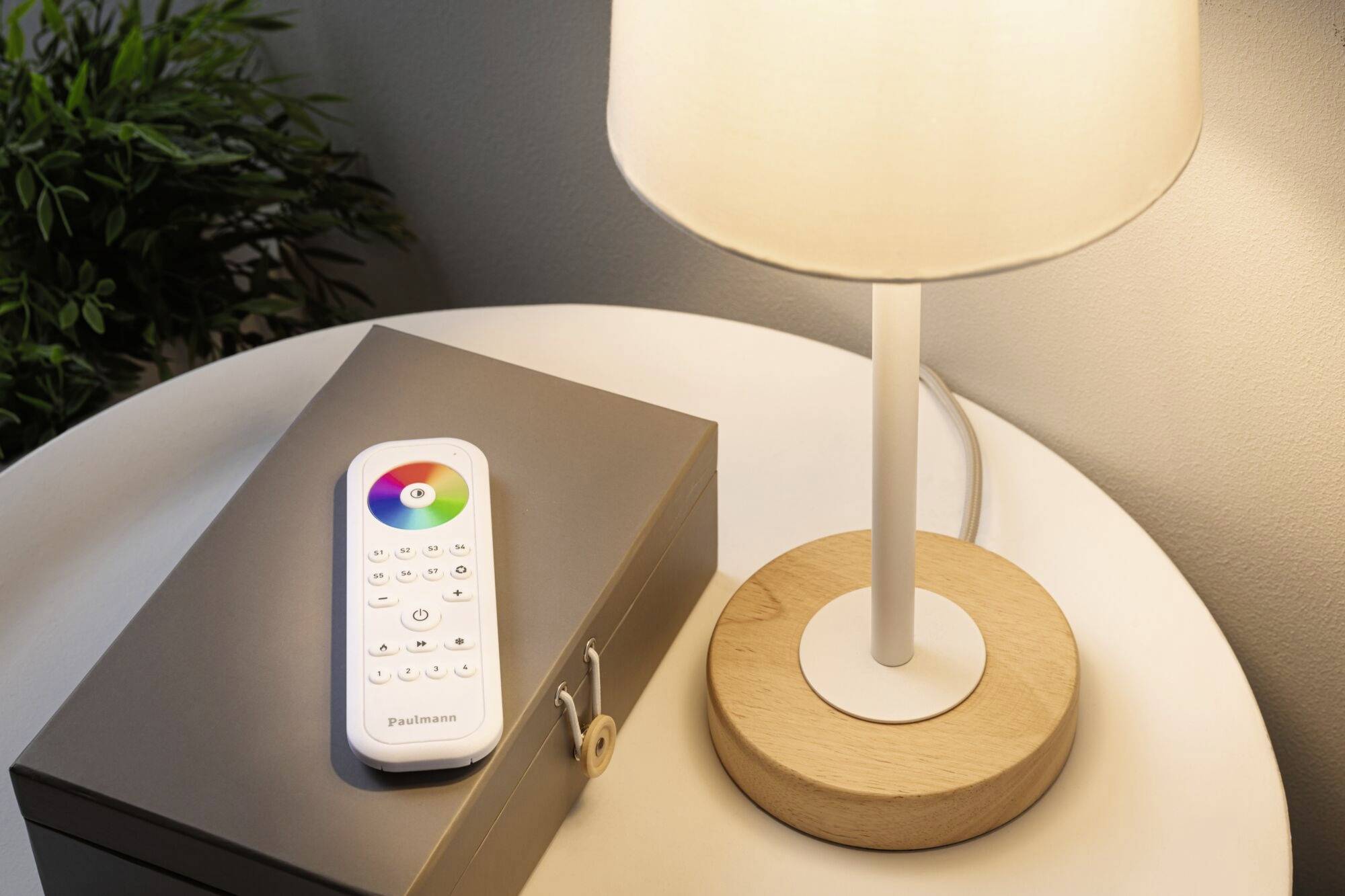 A table lamp with a wooden base illuminates a round table. Next to it lies a remote control with a colour wheel, in front of a grey box.