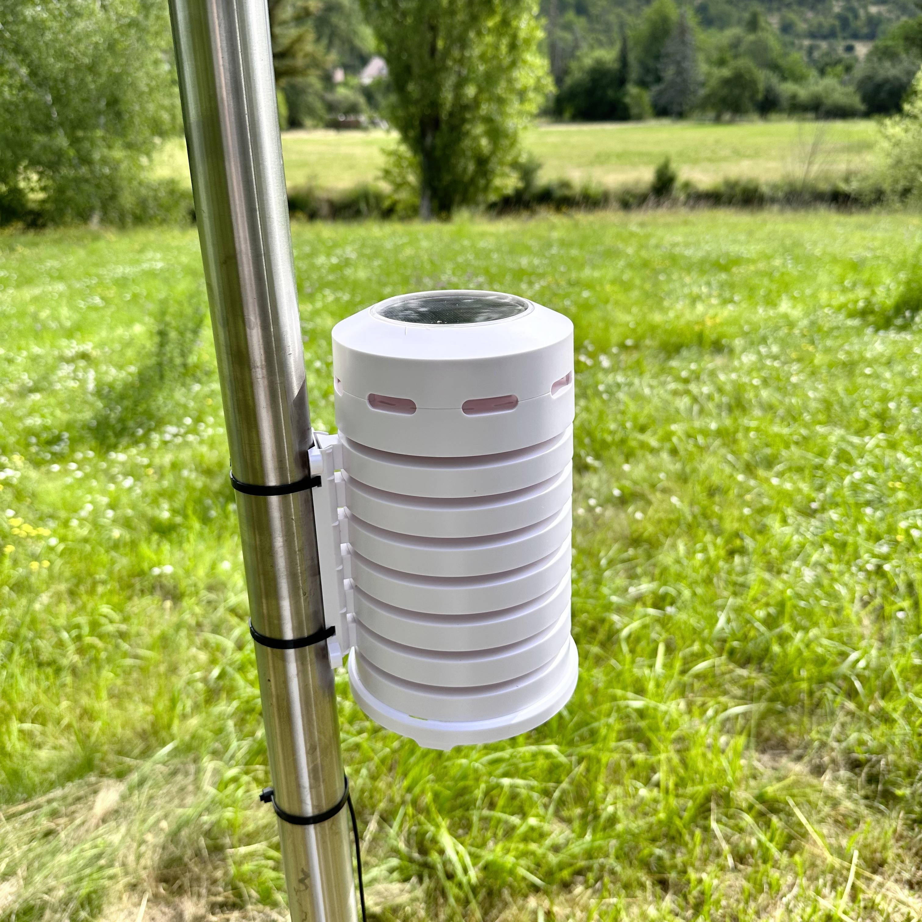 A white humidity and temperature gauge is mounted on a metal pole in a green meadow landscape.