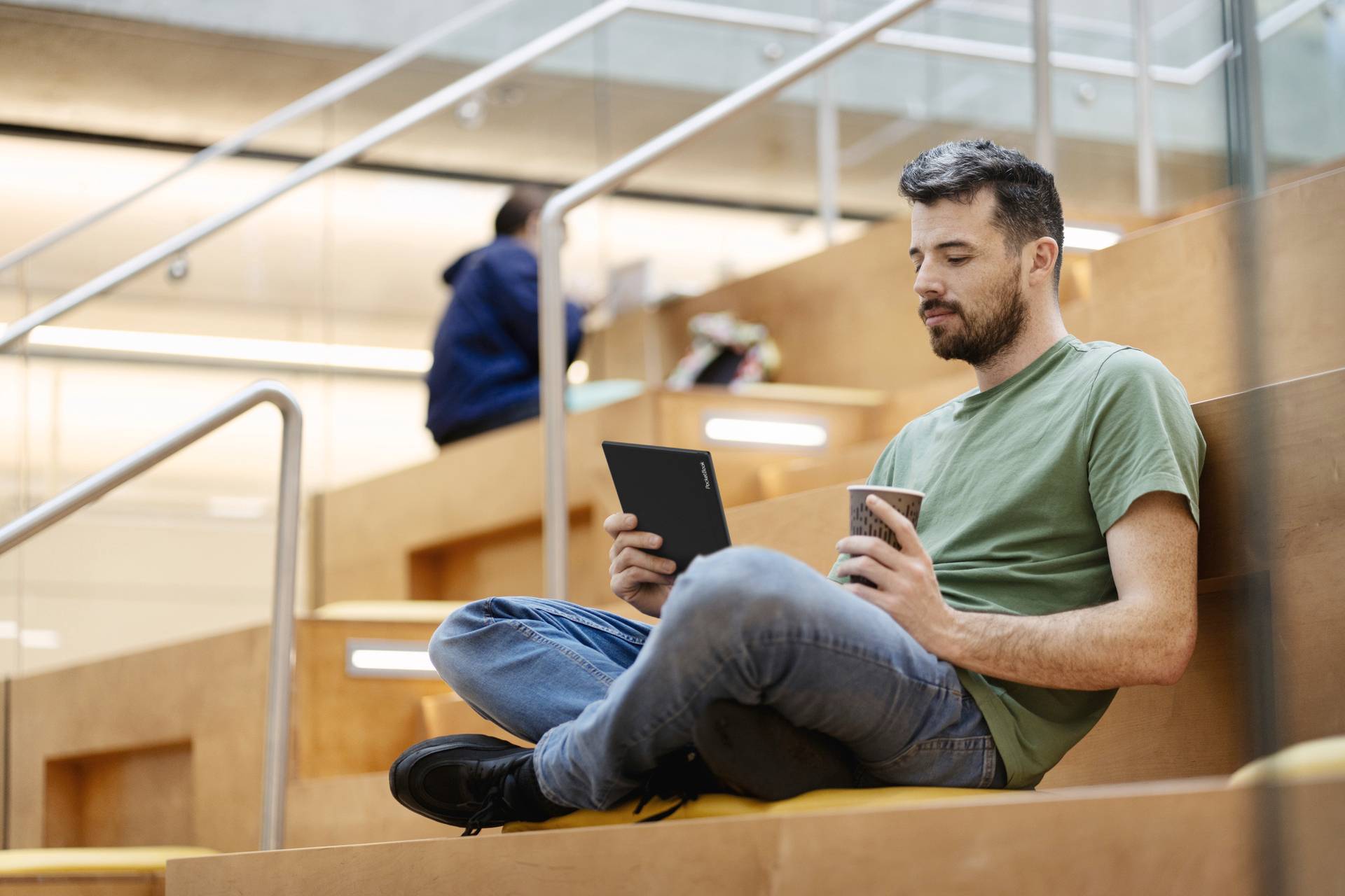 A man is sitting relaxed on a staircase in a modern building, holding a tablet and drinking coffee. Another person is in the background.