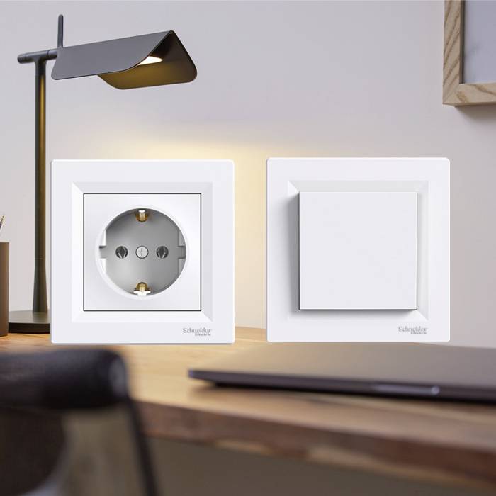 White socket and light switch on a wooden wall next to a modern, black desk lamp. Blurred desk in the background.
