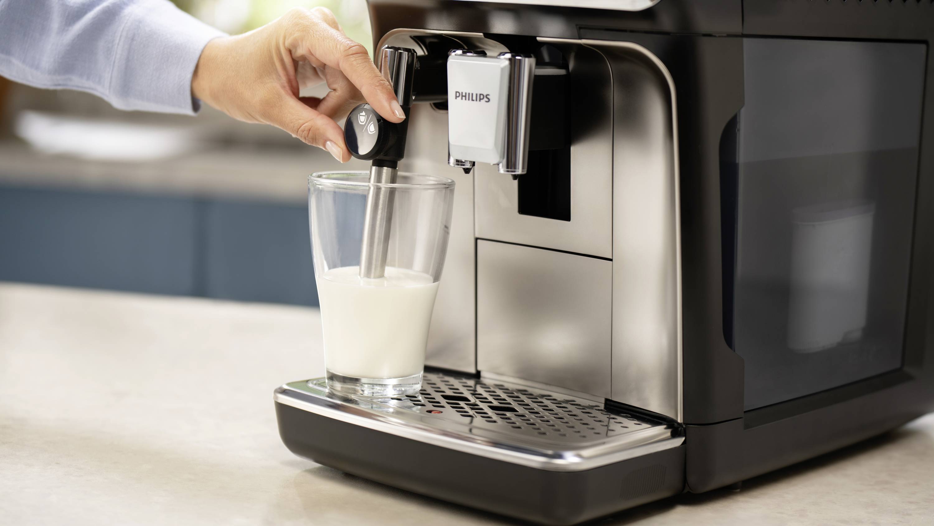 A hand is holding a glass underneath a milk frother on a coffee machine. Milk is being frothed. A kitchen can be seen in the background.