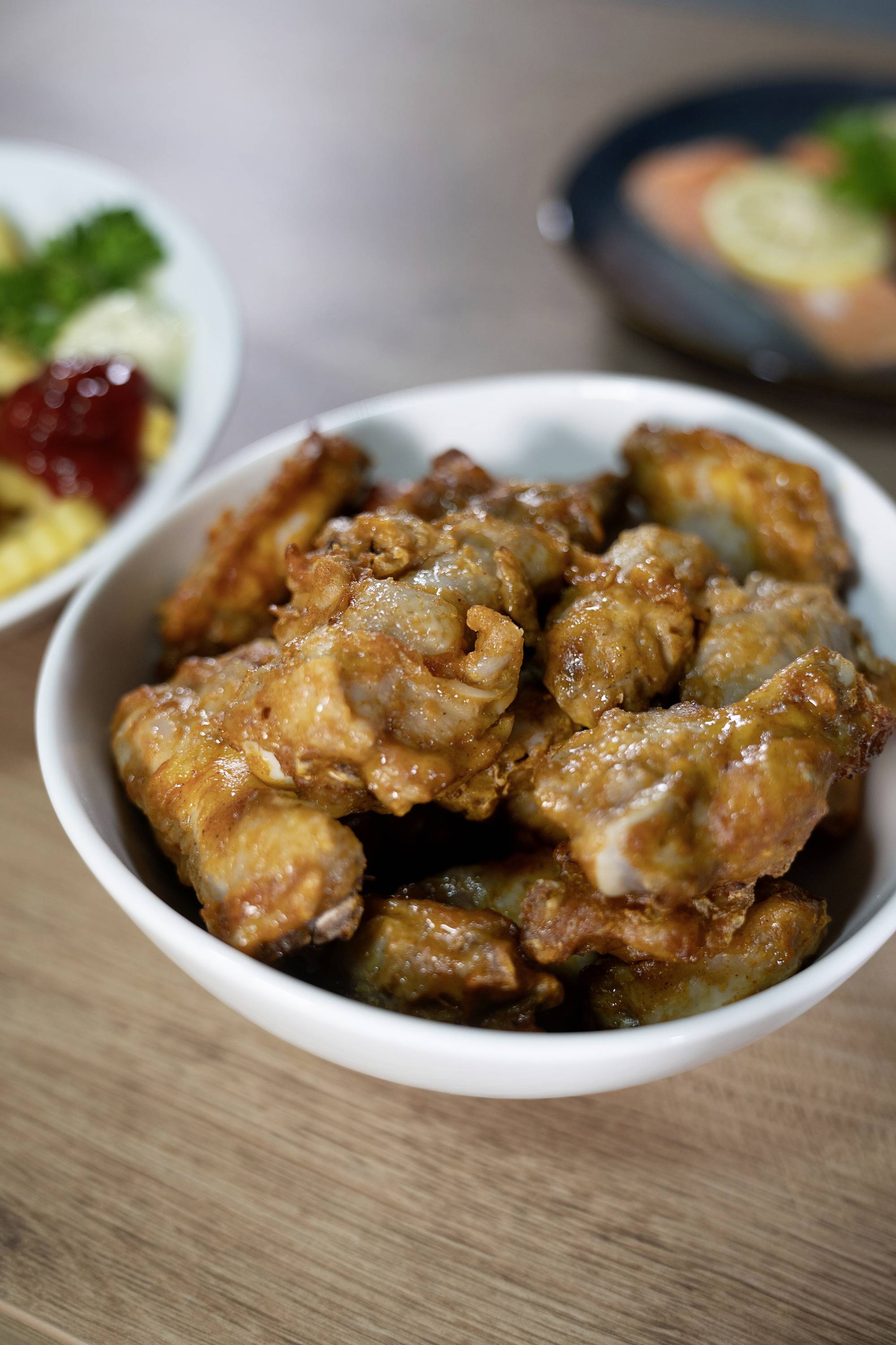 A bowl of fried chicken wings on a wooden table in the foreground, with blurred side dishes in the background.