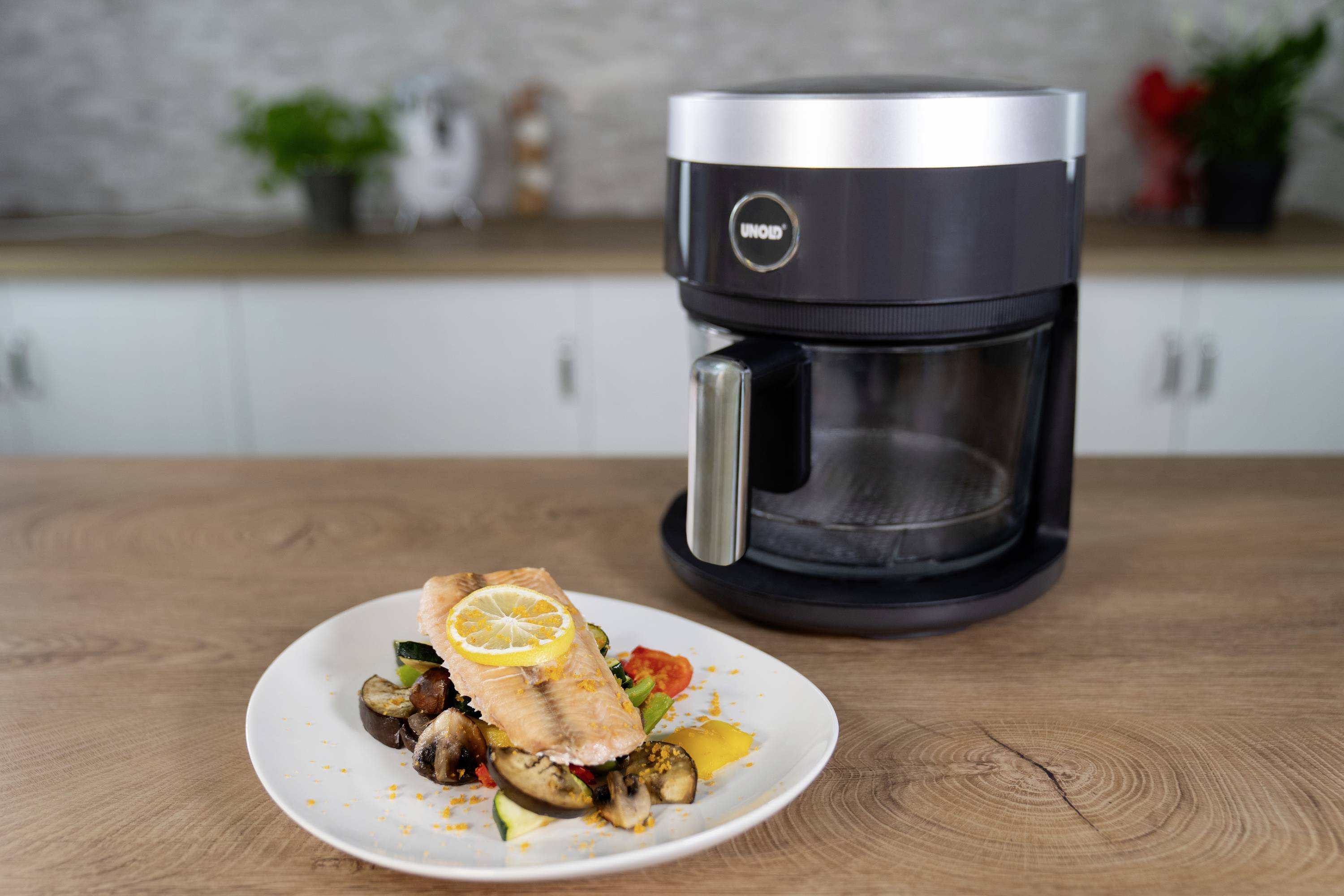 A plate of grilled salmon on vegetables, placed in front of a black food processor on a wooden worktop in a modern kitchen.