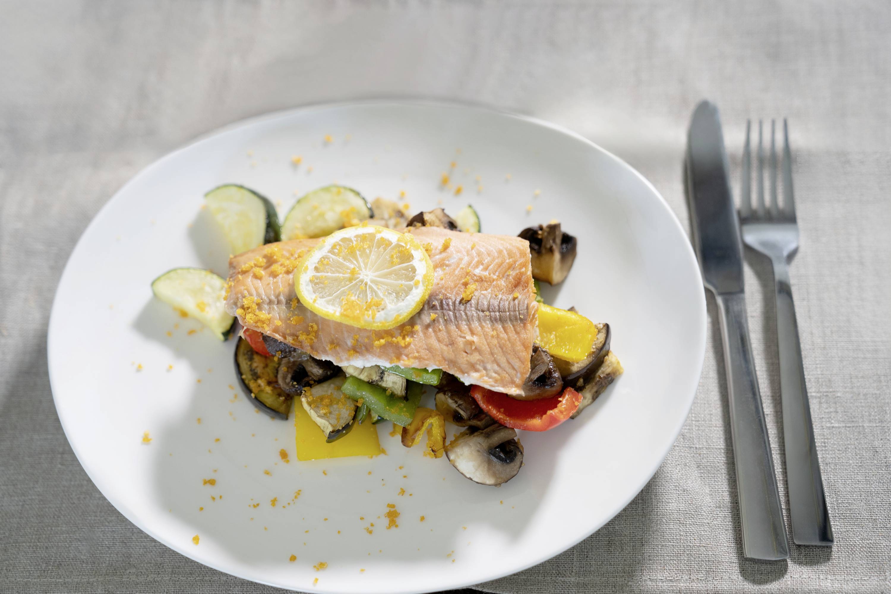 A plate of pan-fried salmon on a bed of courgettes, peppers, tomatoes and mushrooms, garnished with a lemon slice.