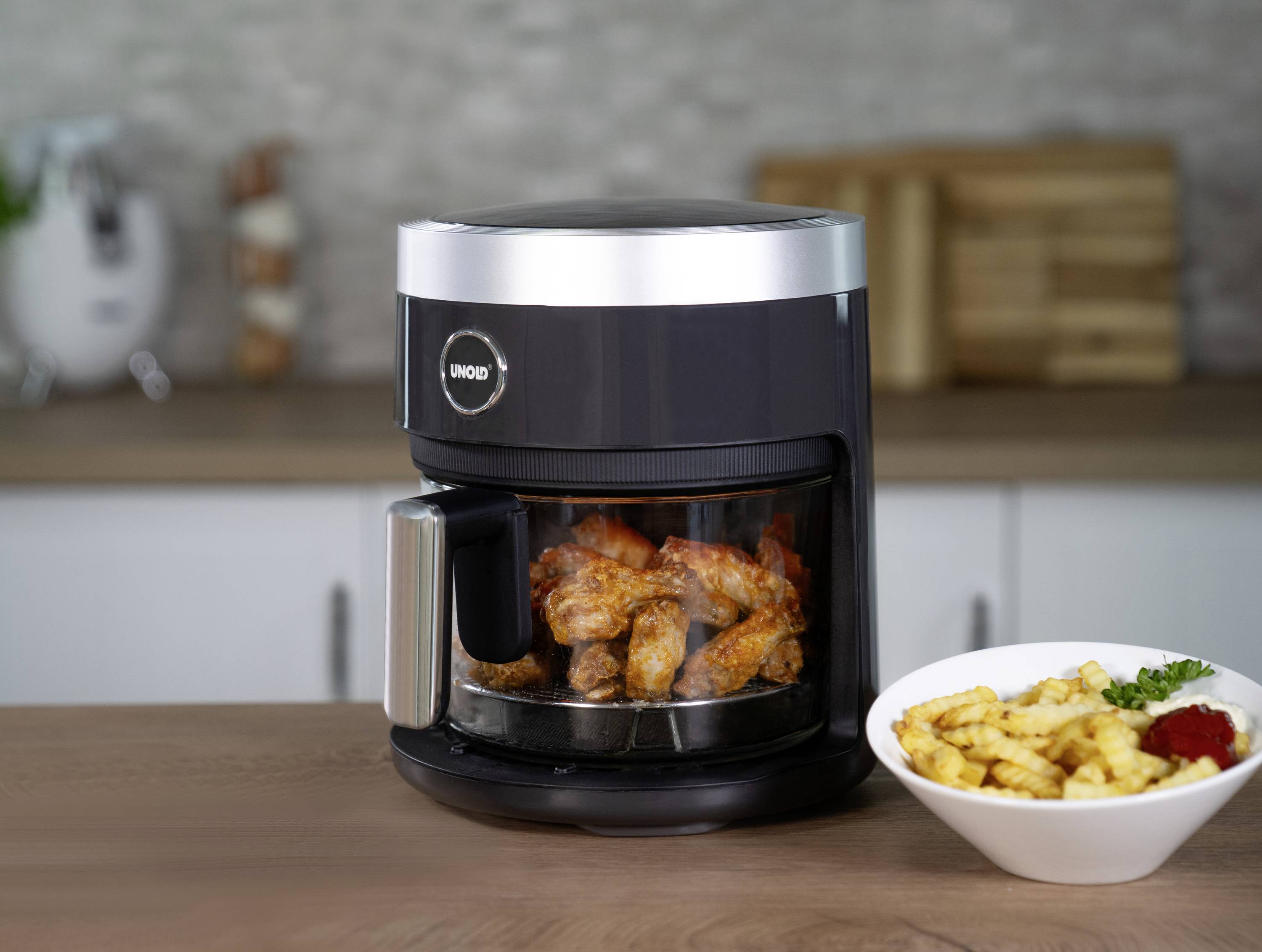 A black air fryer with fried chicken legs on a kitchen worktop. Beside it sits a bowl of chips.