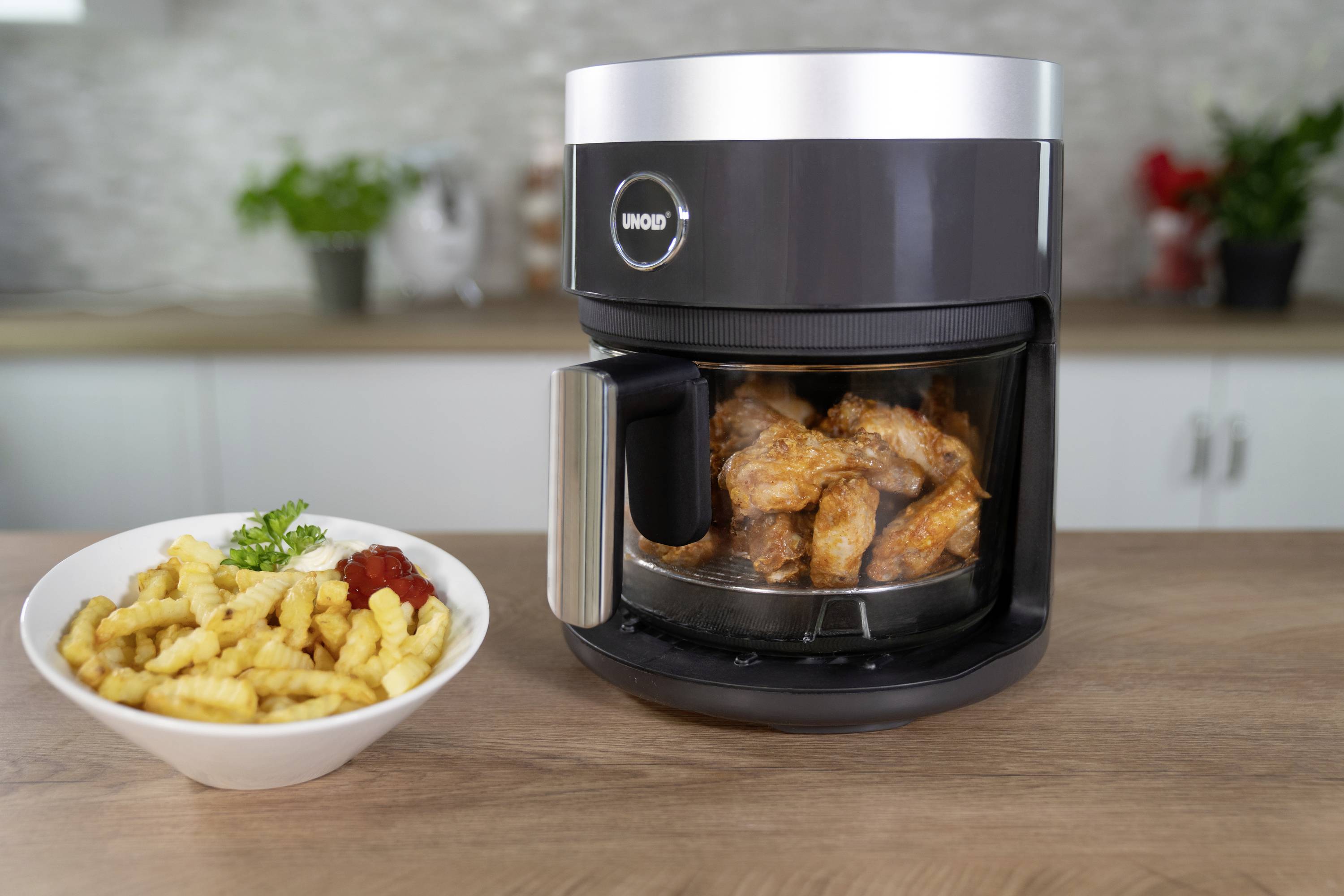 An air fryer filled with chicken wings sits on a kitchen table. Beside it is a bowl of chips with ketchup.