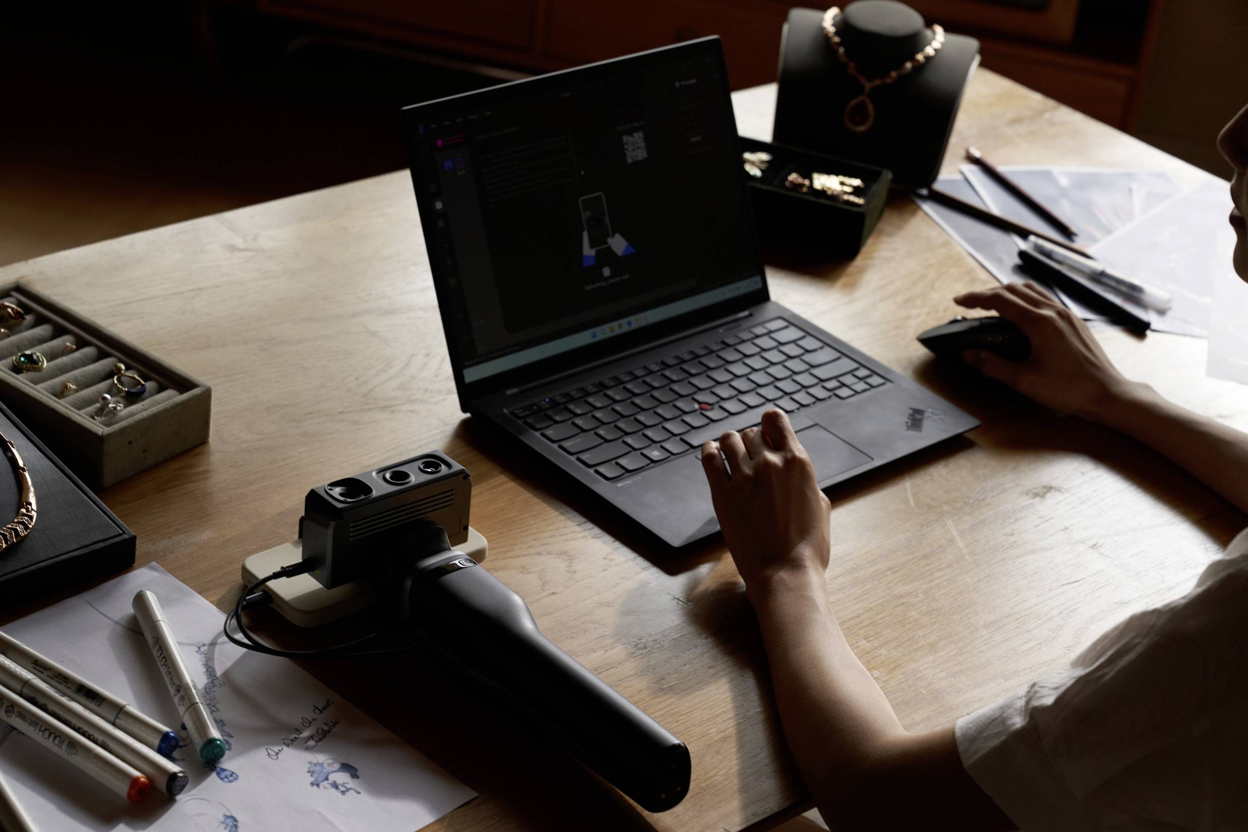 A person is working on a laptop, surrounded by jewellery pieces and sketches. A 3D scanner is also visible on the table.