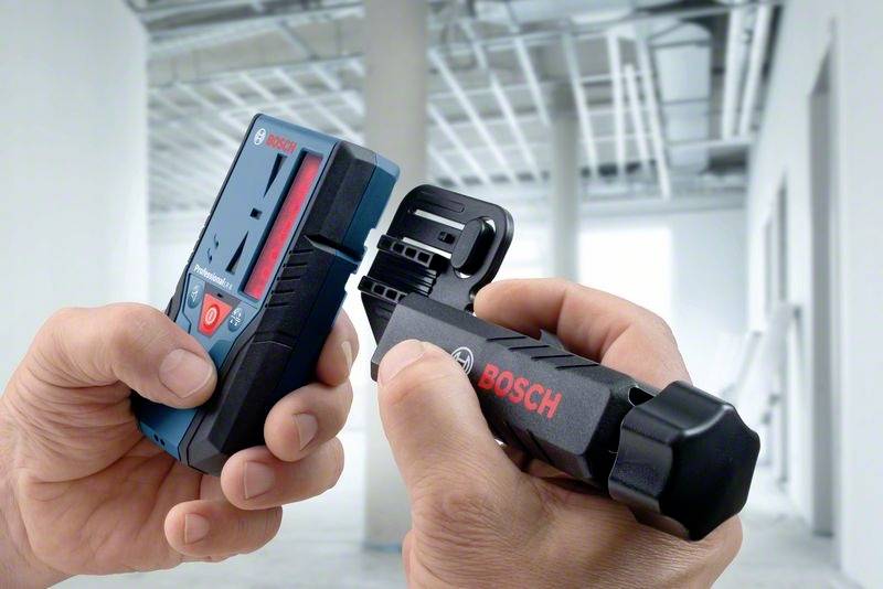 Two hands are holding a blue and a black Bosch device in front of a blurred, modern interior space with visible pipework.