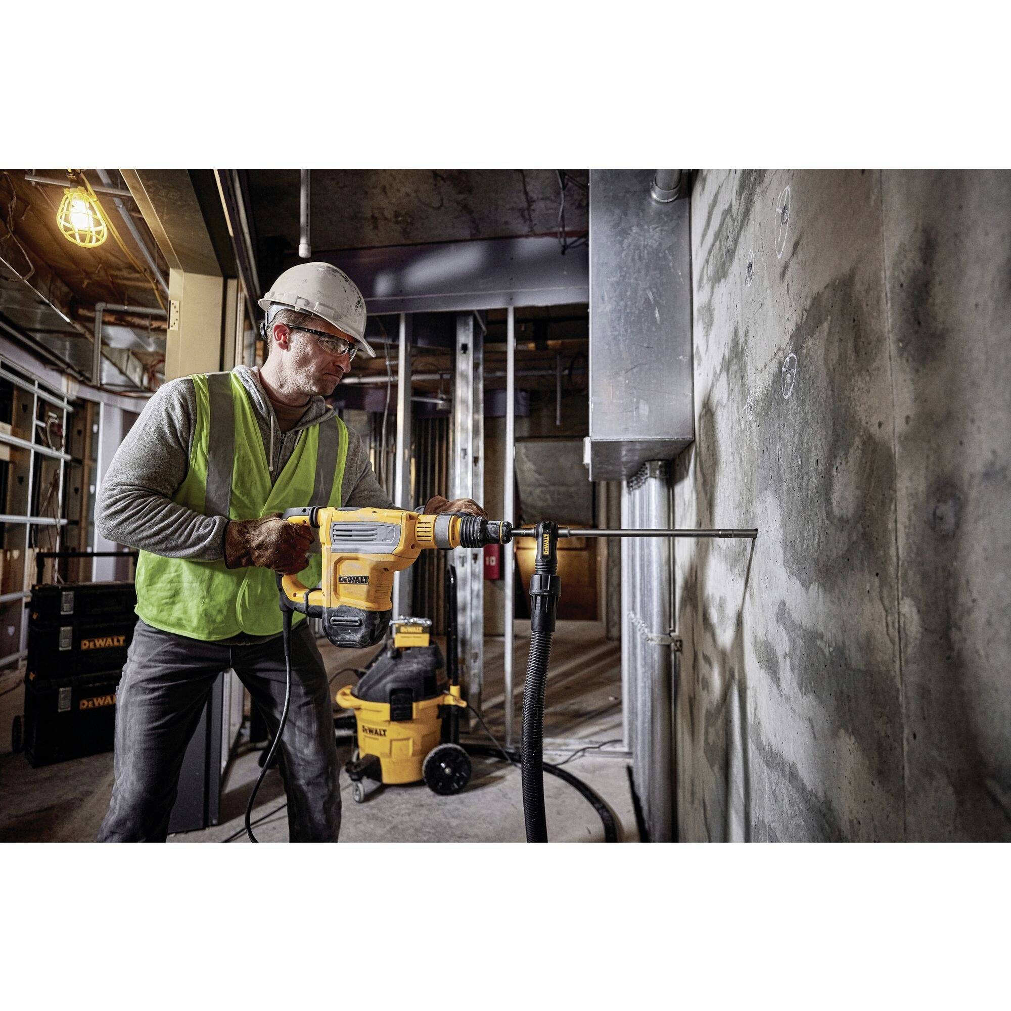 A worker in safety clothing is drilling into a concrete wall on a building site, surrounded by scaffolding and tools.