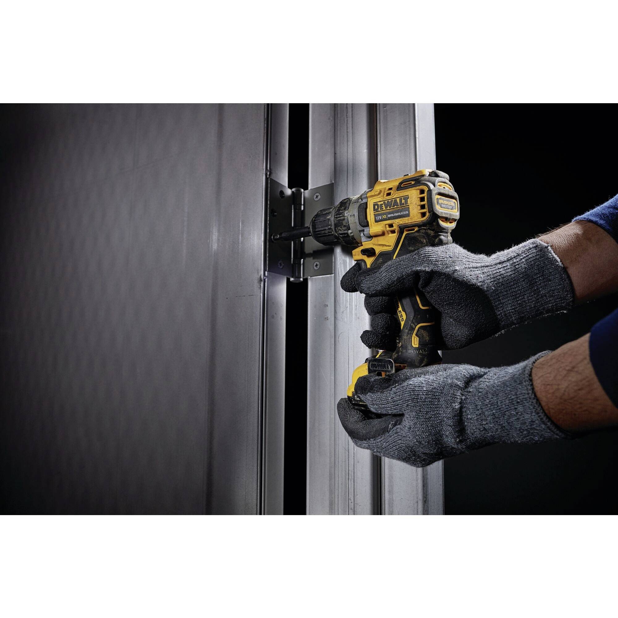 A person wearing grey gloves is holding a yellow cordless drill and tightening a screw in a metal hinge.